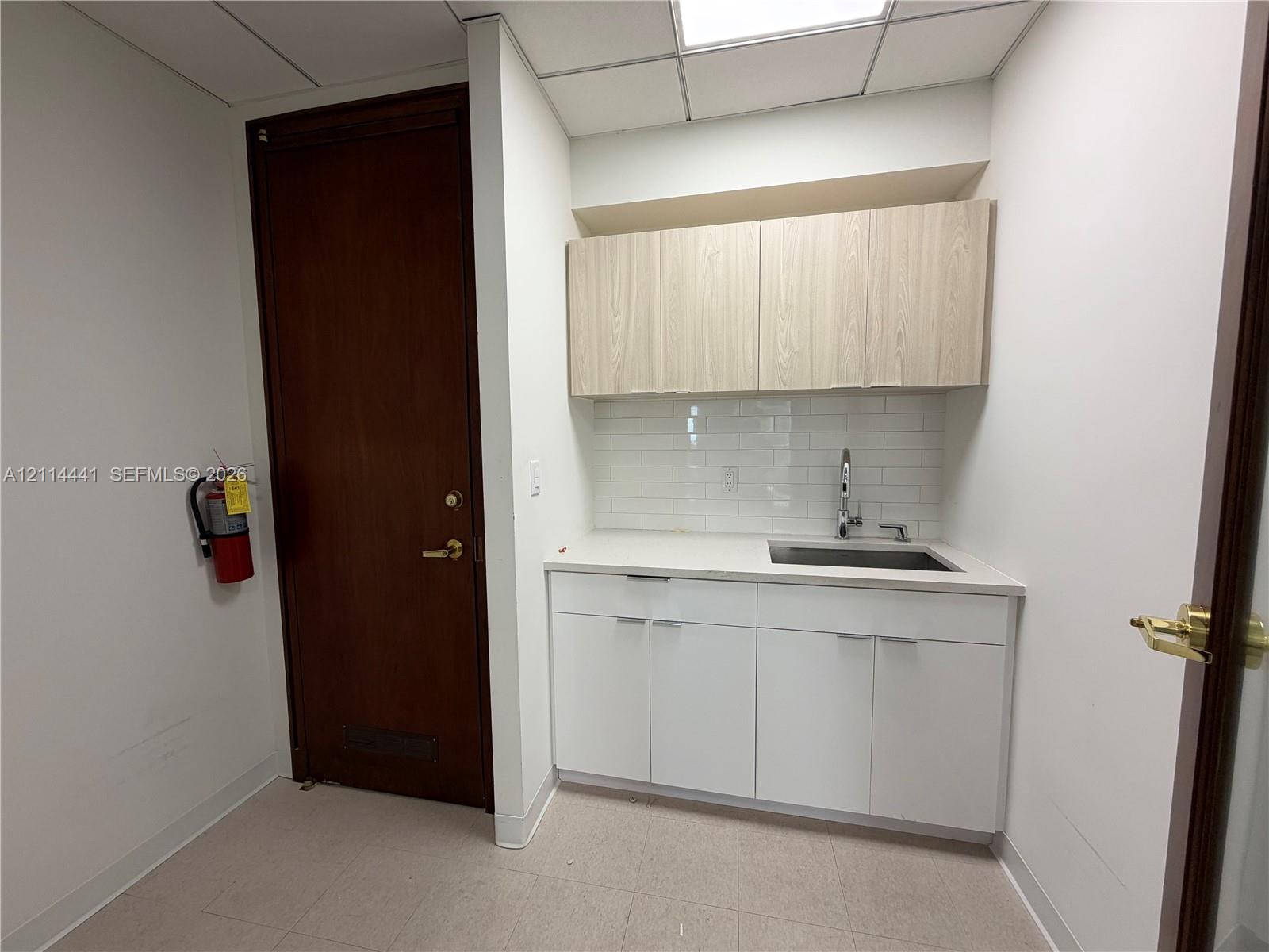 Kitchenette area featuring white cabinetry, wood-finish upper cabinets, solid-surface countertops, a tiled backsplash, and an integrated sink