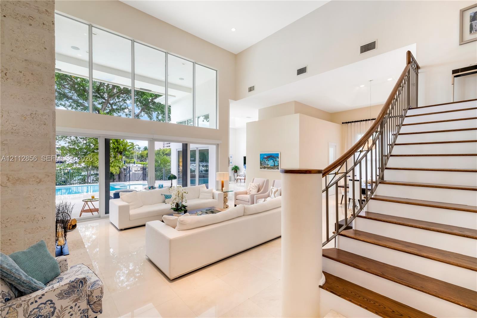 Double-height ceilings, floor-to-ceiling glass windows, staircase with wood-finish treads and metal railings, stone-faced interior column, and light-t