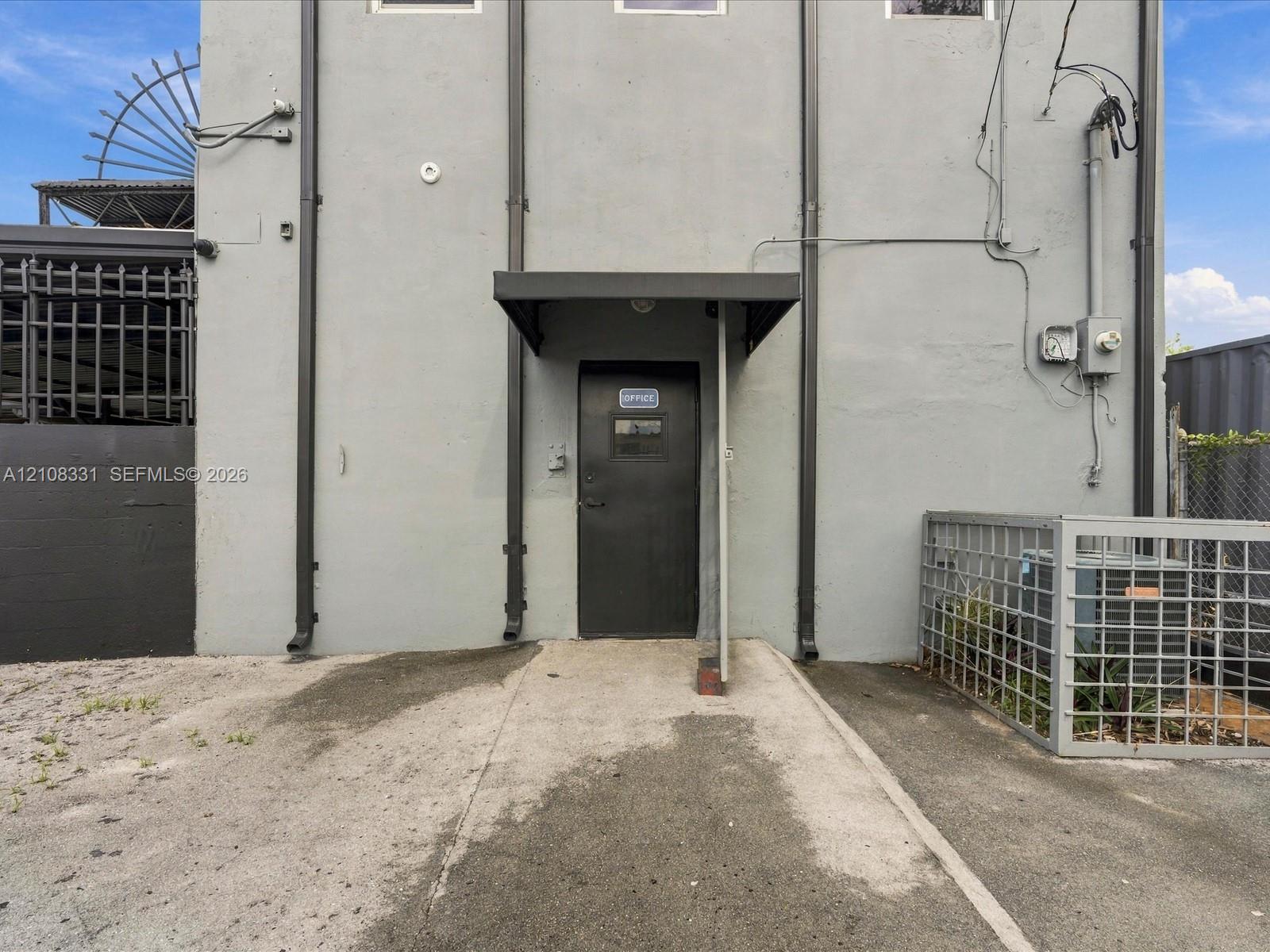 Exterior entry featuring a dark-toned steel door with an overhead canopy, concrete surfacing, and metal perimeter fencing