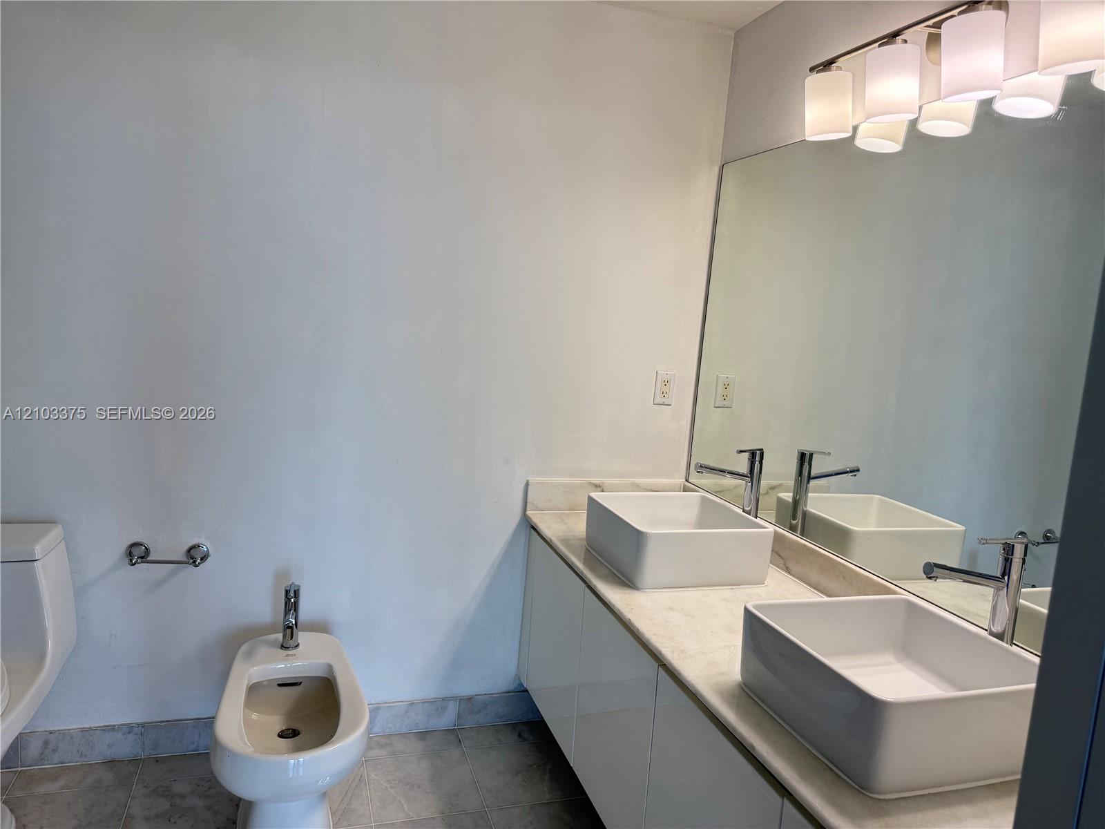 Dual-sink vanity with white basin vessel sinks, light-toned stone countertop, white cabinetry, wall-to-wall mirror, and overhead multi-light fixture