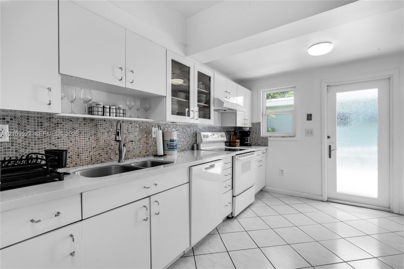 Kitchen featuring white cabinetry with glass-front display uppers, mosaic tile backsplash, solid-surface countertops, dual-basin stainless steel sink 