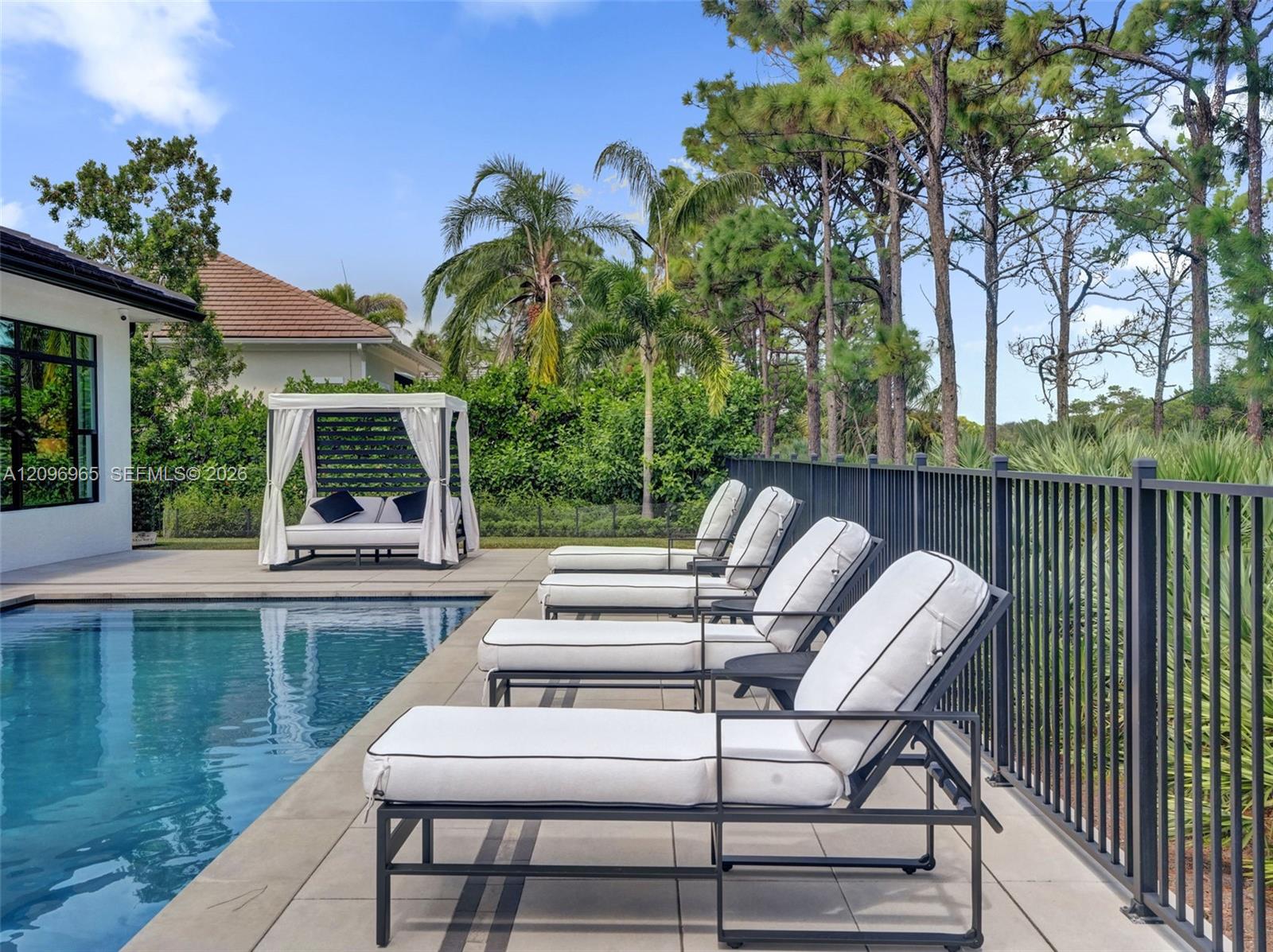 In-ground swimming pool with stone-tiled decking and black metal perimeter fencing, featuring mature pine tree landscaping