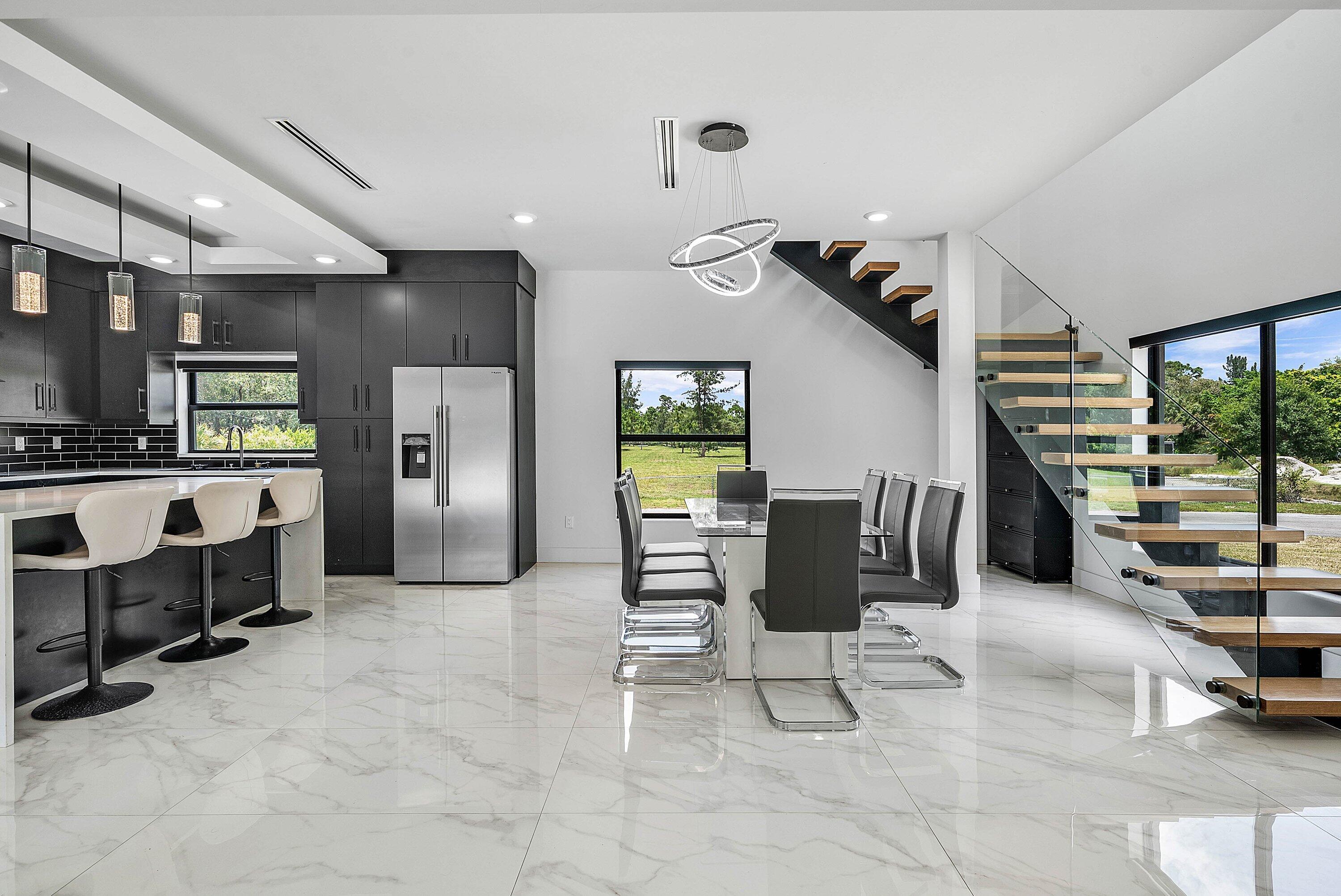 Open-concept interior featuring large format marble-look tile flooring, a modern floating staircase with glass railing, a kitchen with dark cabinetry
