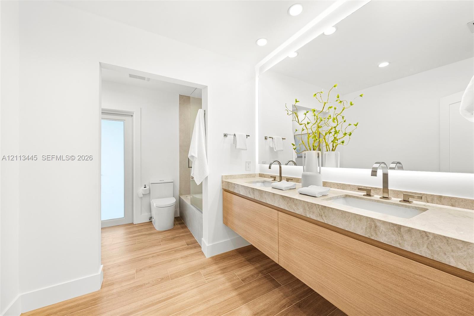 Floating dual vanity with stone countertop, under-mount sinks, modern metal faucets, wood-finish cabinetry, and oversized wall-mounted LED mirror