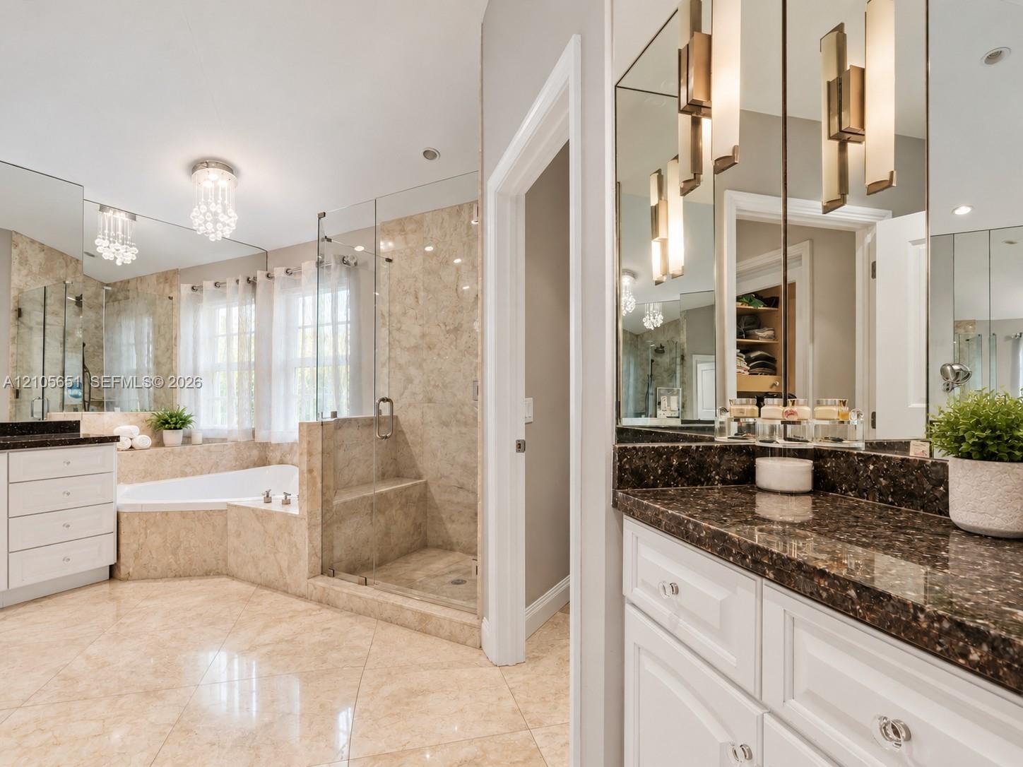 Bathroom featuring stone-finish flooring and wall tiling, dual white vanities with dark stone countertops, a frameless glass-enclosed shower with a bu