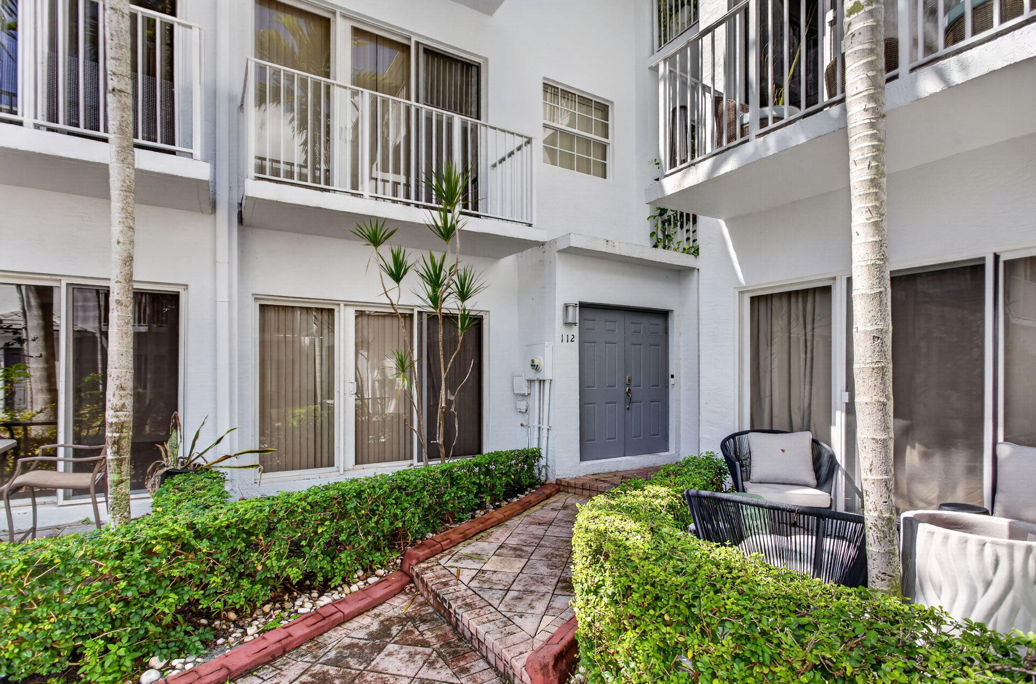 White stucco exterior with a gray double entry door, multiple sliding glass doors, upper-level balconies with white railings, and a tiled walkway