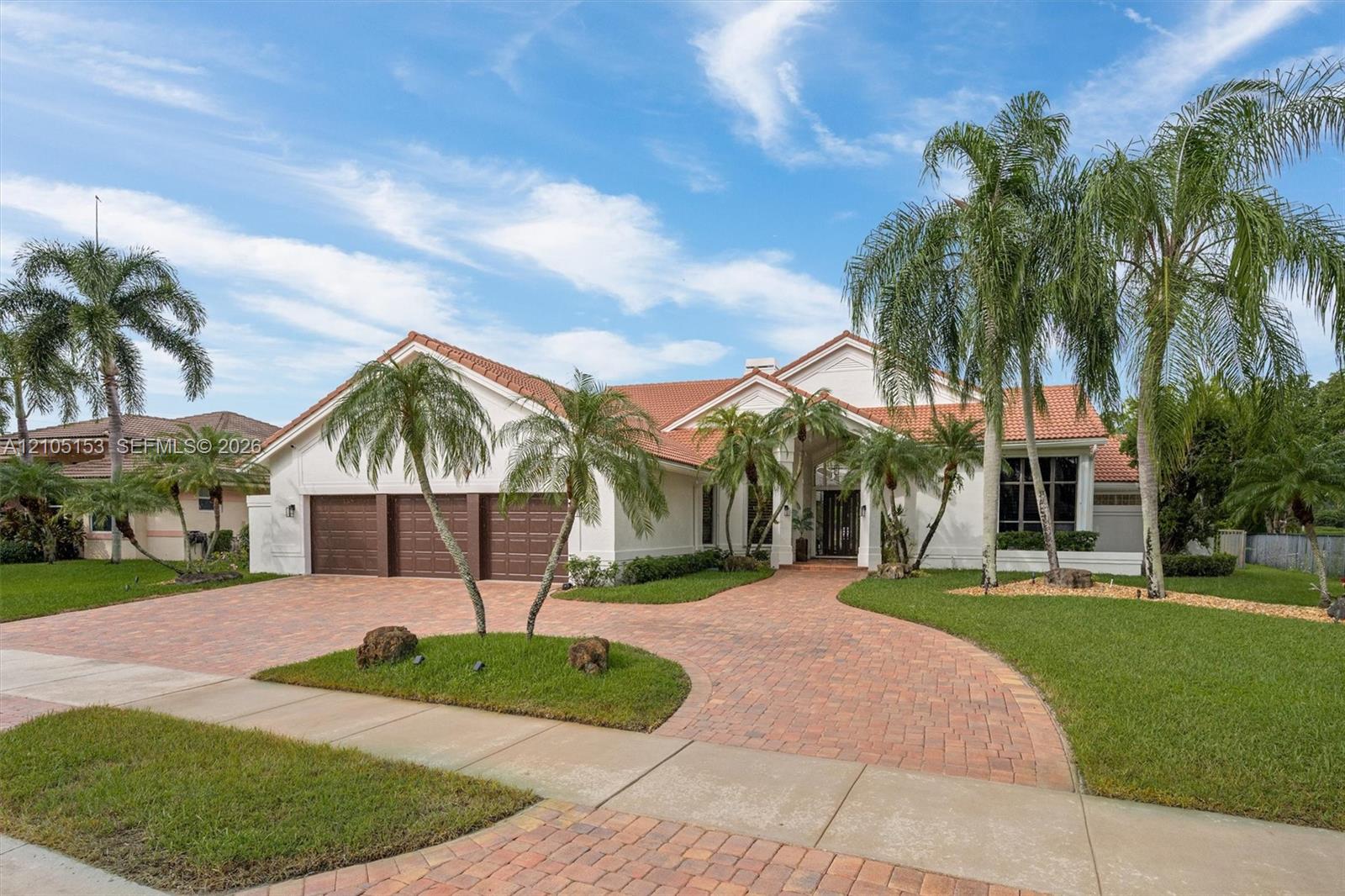White stucco exterior with a terra cotta tile roof, a curved paver driveway, a two-car garage with wood-look doors, and lush landscaping