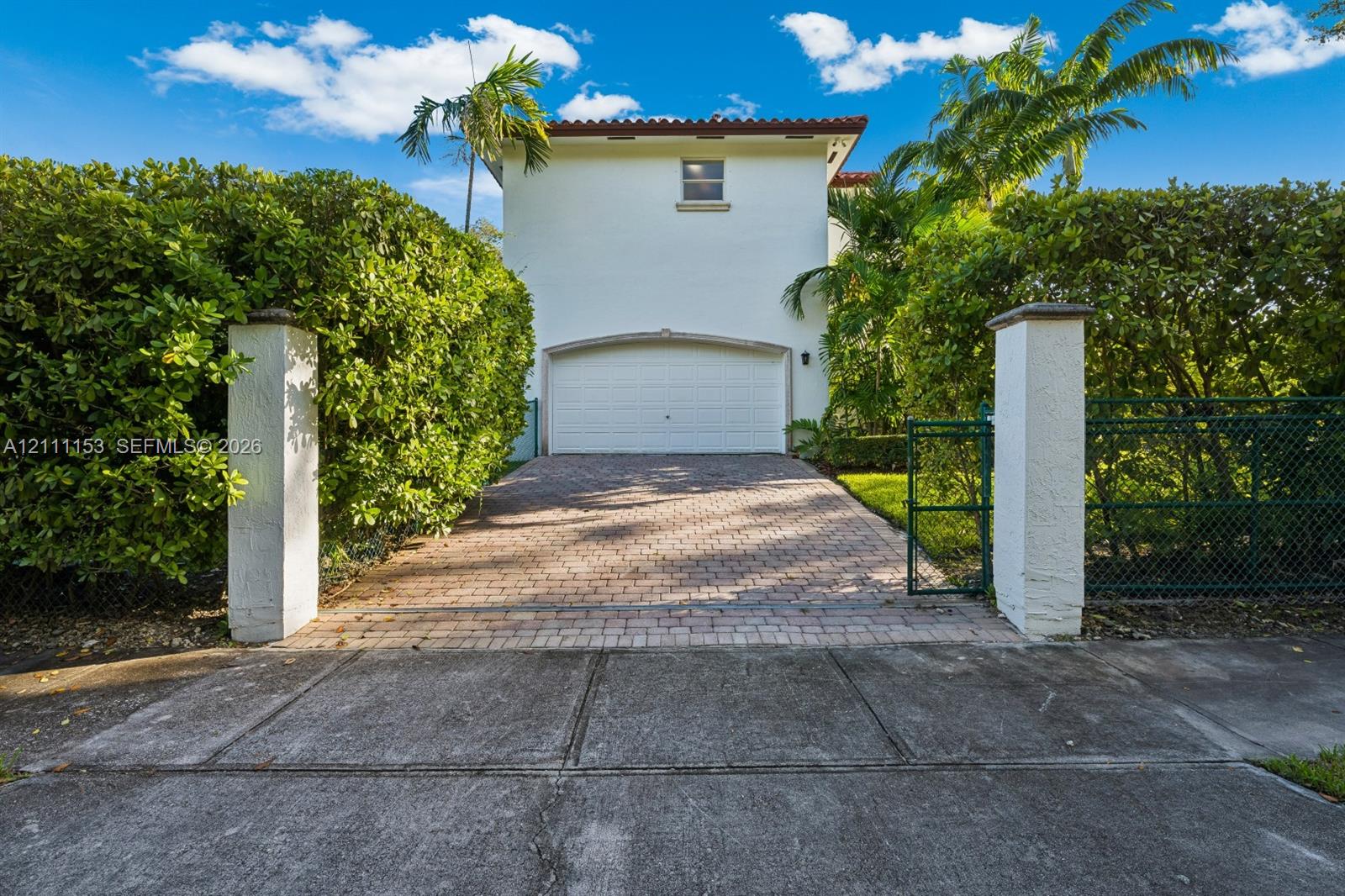 Stucco exterior featuring a paver driveway, arched garage entry, decorative stucco pillars, and mature landscaping