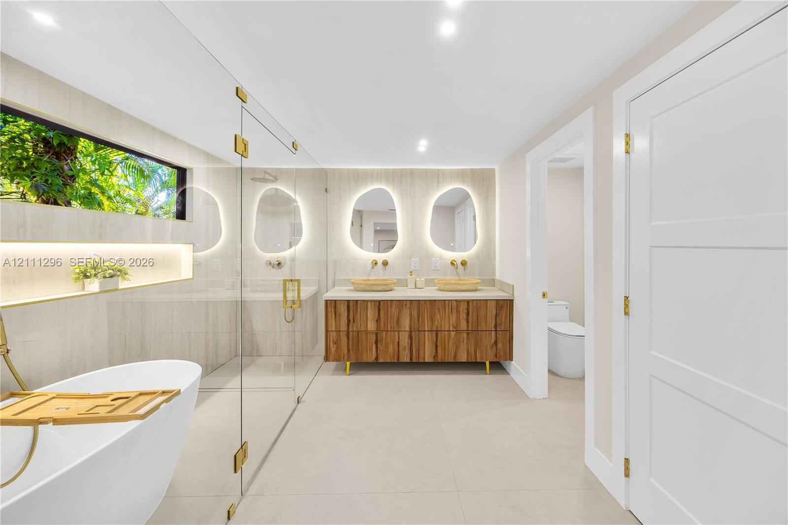 Freestanding soaking tub with gold-tone fixtures, wood-finish floating dual vanity, stone-topped surfaces, wall-mounted faucets, and back-lit organic-