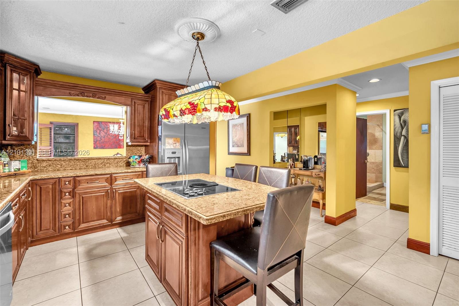 Kitchen featuring wood-finish cabinetry, light-toned tile flooring, and a center island with a built-in cooktop and stone-look countertop