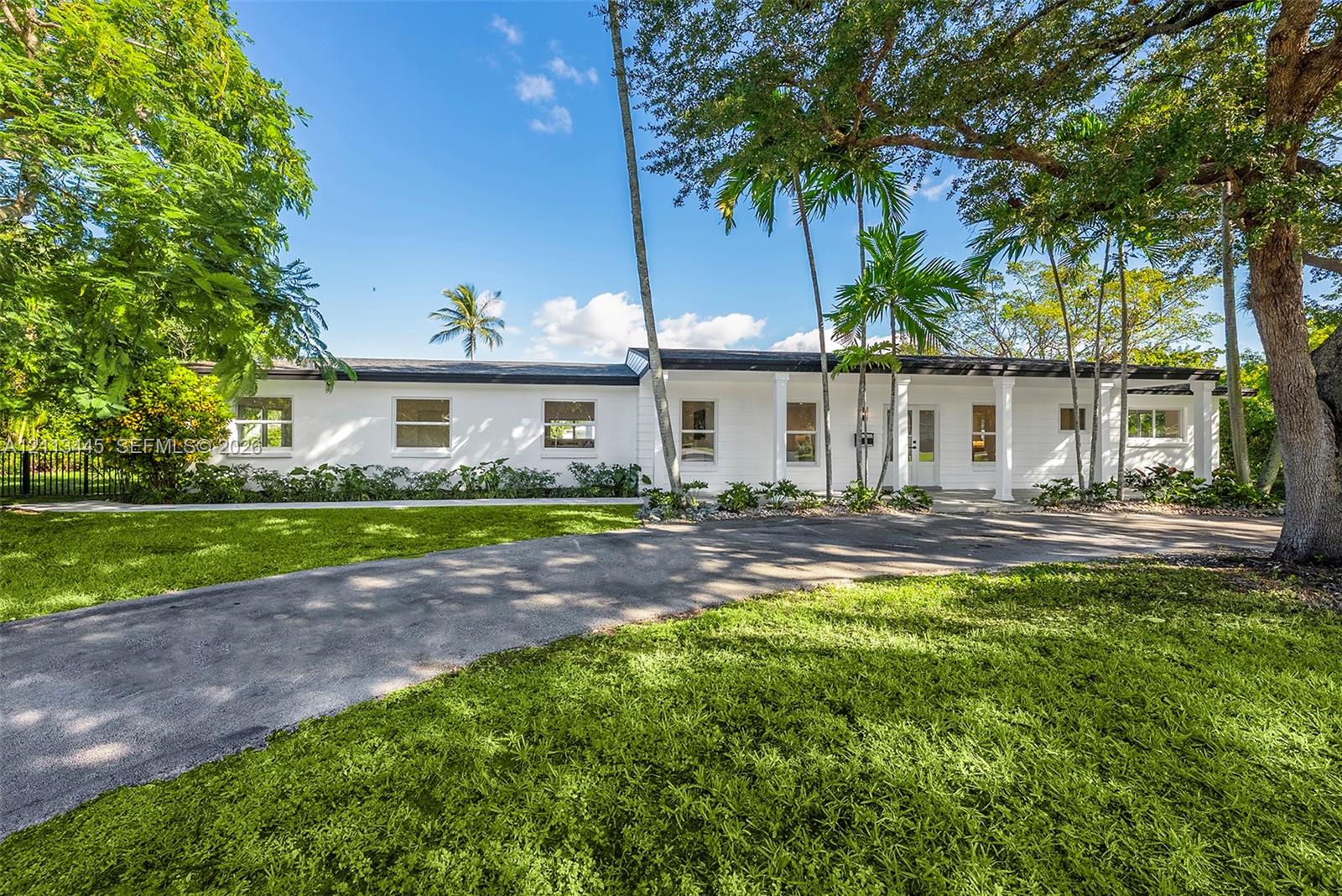 Single-story exterior featuring white masonry walls, a covered front portico with columns, a sprawling lawn, a paved driveway, and mature palm trees