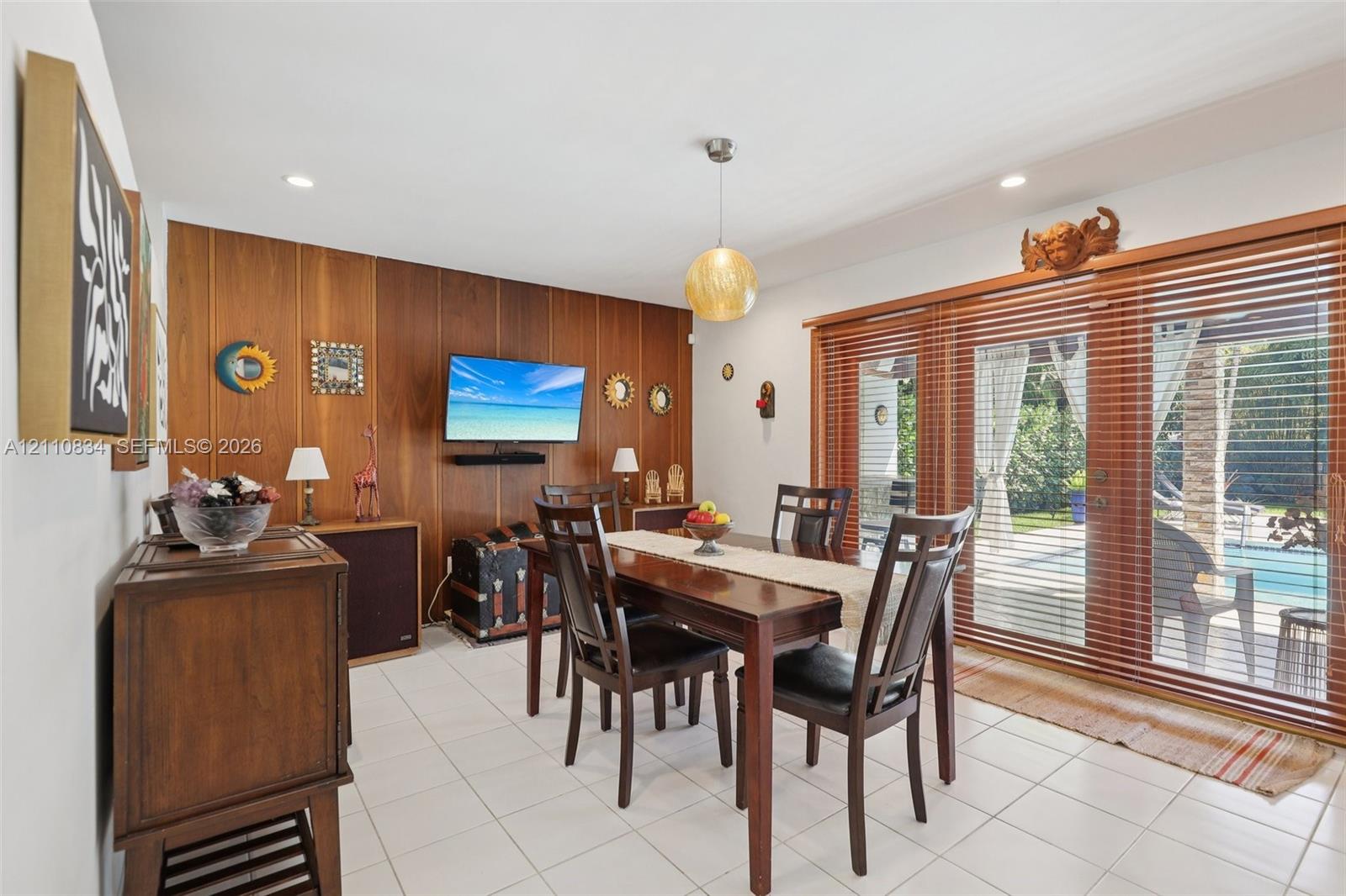 Dining area featuring white tile flooring, wood-paneled accent wall, recessed lighting, spherical pendant light, and sliding glass doors with wood bli