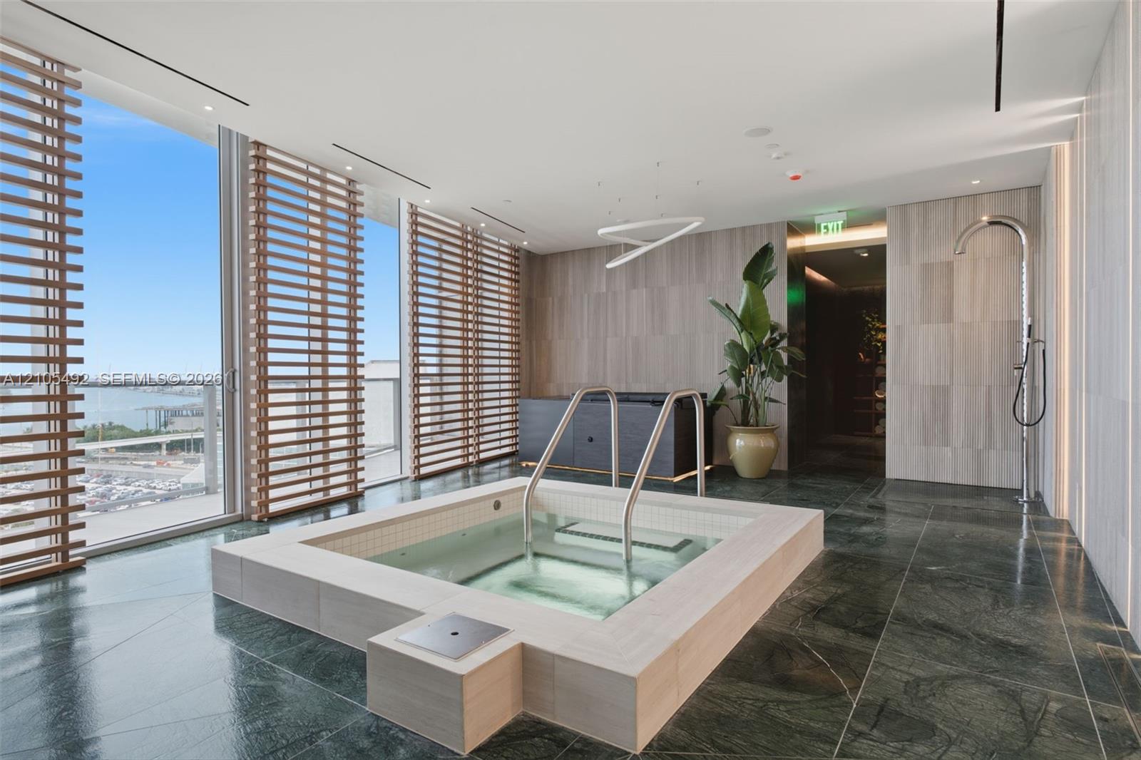 Sunken jetted tub with integrated handrails, floor-to-ceiling glass paneling, wood-slat privacy screening, polished stone flooring, and wall-mounted s