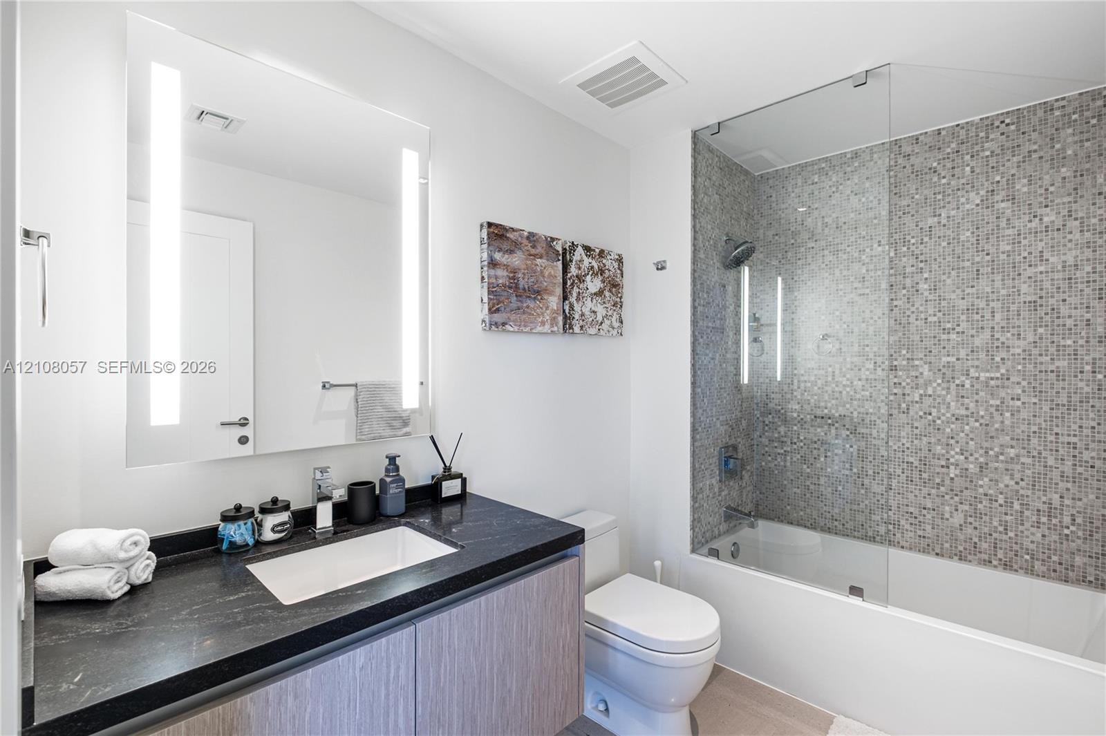Vanity with a dark countertop and integrated rectangular sink, wood-finish cabinetry, illuminated mirror, and a tiled shower-tub combination with a gl