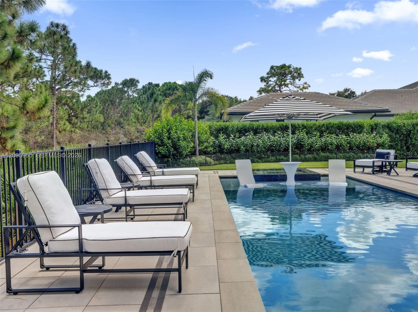 Outdoor swimming pool with built-in tanning ledge, raised spillway spa, neutral-toned paved pool deck, and metal perimeter fencing