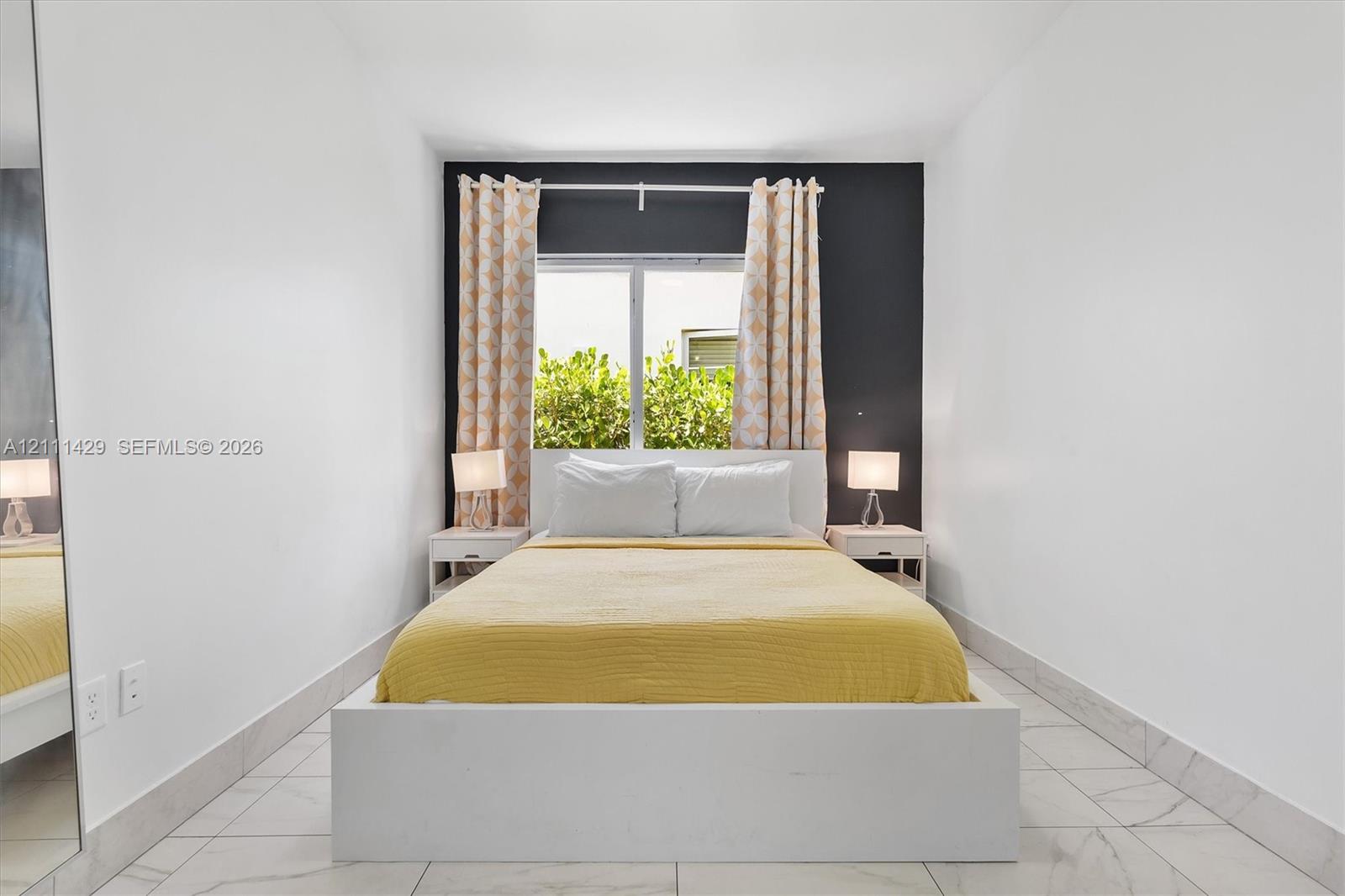 Bedroom interior featuring large-format tile flooring, a dark charcoal accent wall, a central window, and an integrated floor-to-ceiling mirror