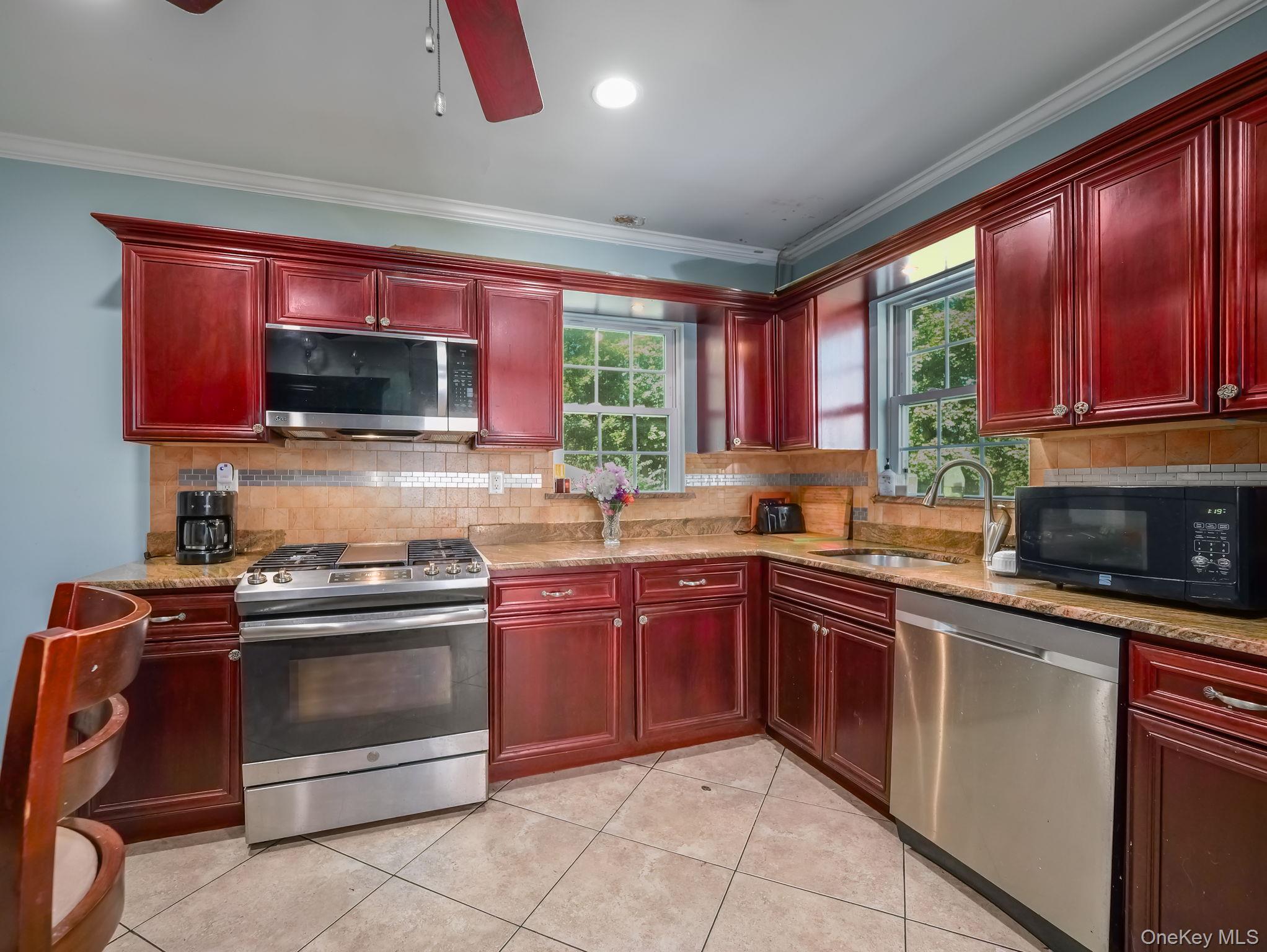 Kitchen with reddish brown cabinets, appliances with stainless steel finishes, light stone counters, decorative backsplash, and crown molding
