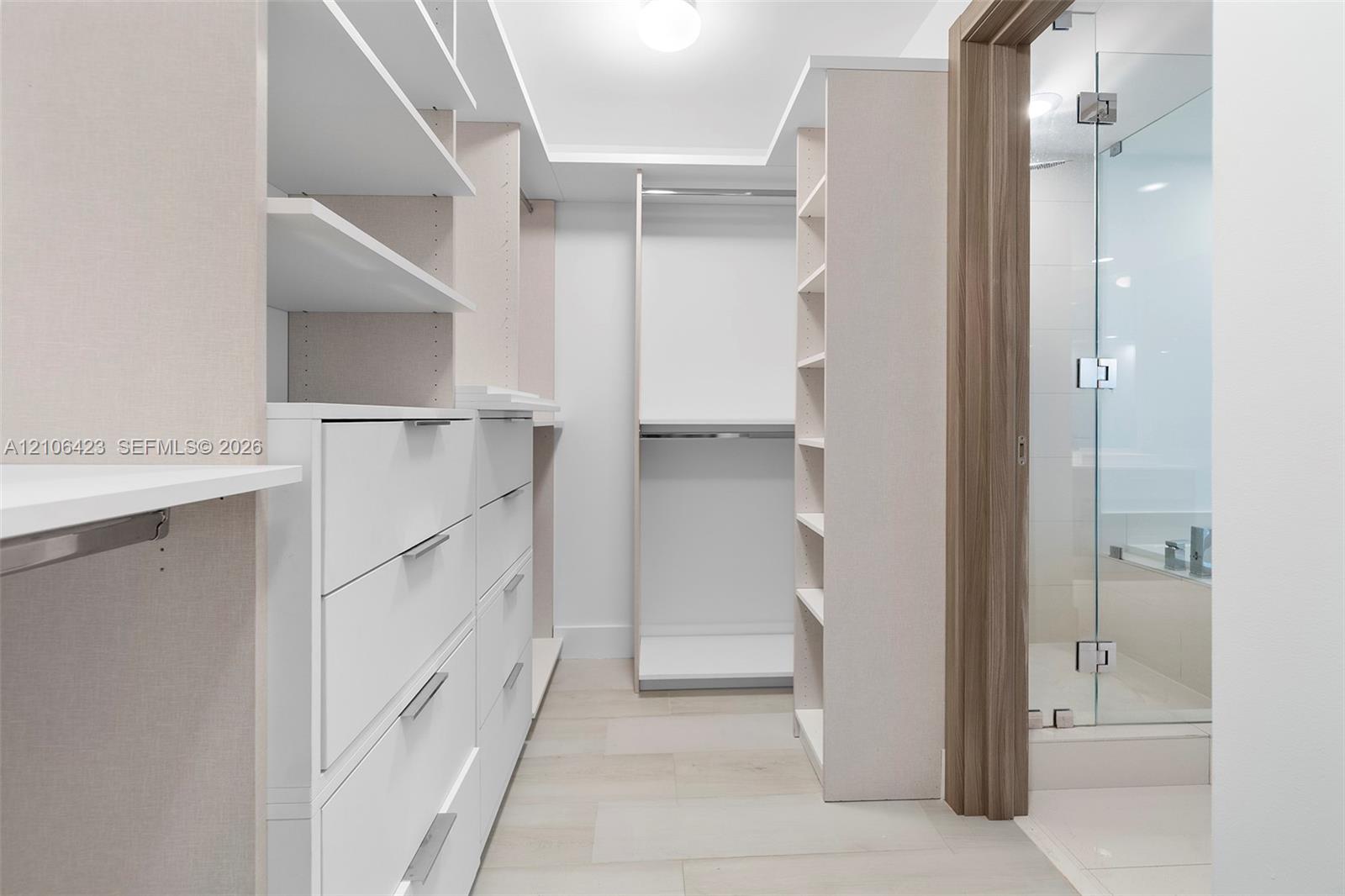 Walk-in closet featuring custom white cabinetry, open shelving, integrated drawer units, and tile flooring, including a glass-enclosed shower with woo