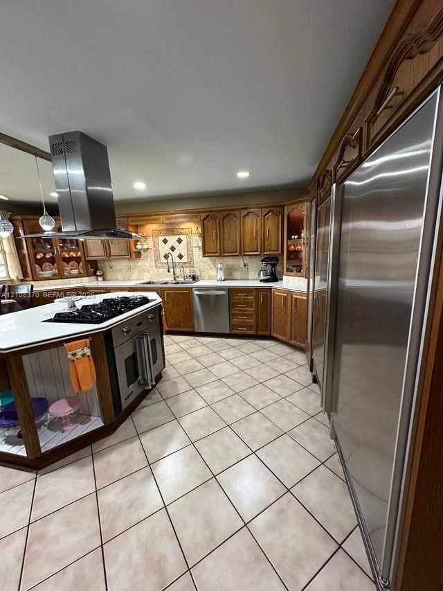 Kitchen featuring wood cabinetry, stainless steel appliances, central island with a built-in cooktop, chimney-style range hood, and tile flooring