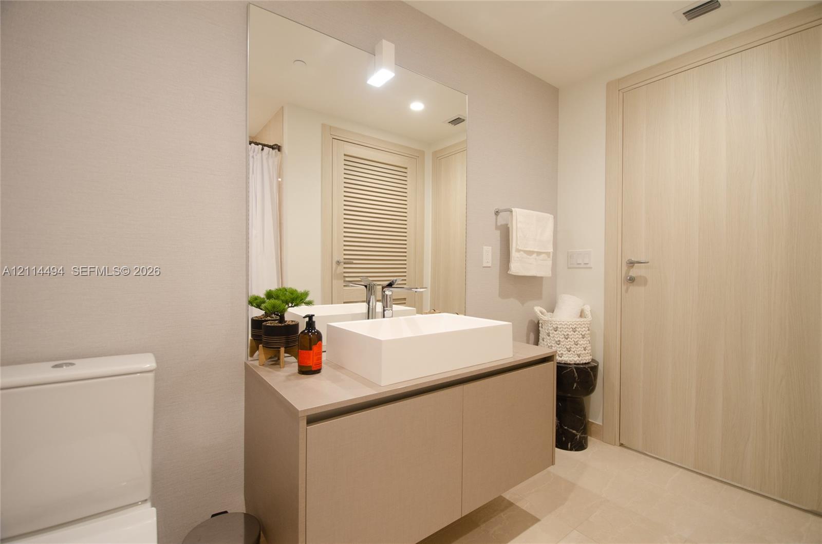 Modern floating vanity with a rectangular vessel sink and wall-mounted faucet, oversized wall mirror, light-toned wood-finish door, and neutral tile f