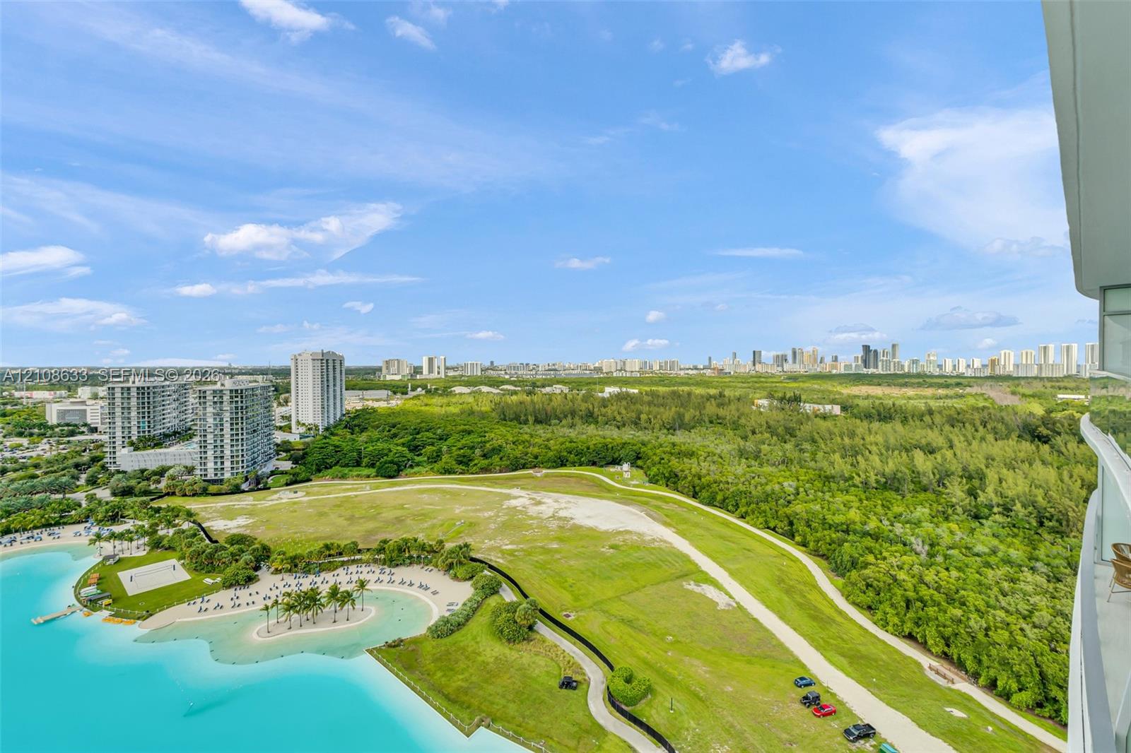 Panoramic outdoor scenery featuring a turquoise lagoon, expansive green space, mature tree canopy, and a distant city skyline