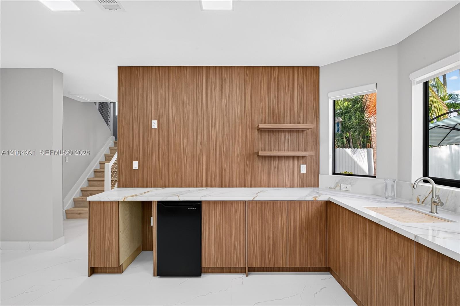 Custom wet bar featuring wood-veneer cabinetry, stone-finish countertops, floating shelving, an undermount sink with a gooseneck faucet, and integrate