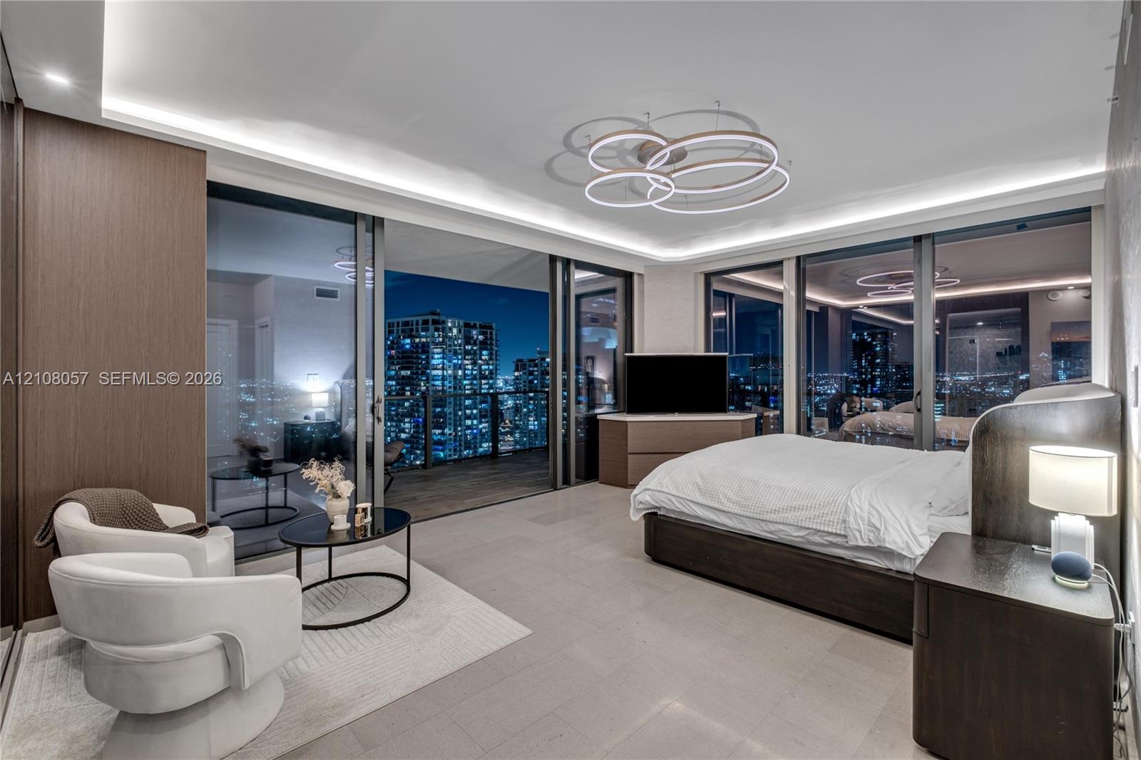 Floor-to-ceiling glass sliding doors, recessed cove lighting, sculptural multi-ring chandelier, large-format tile flooring, and integrated wood-veneer