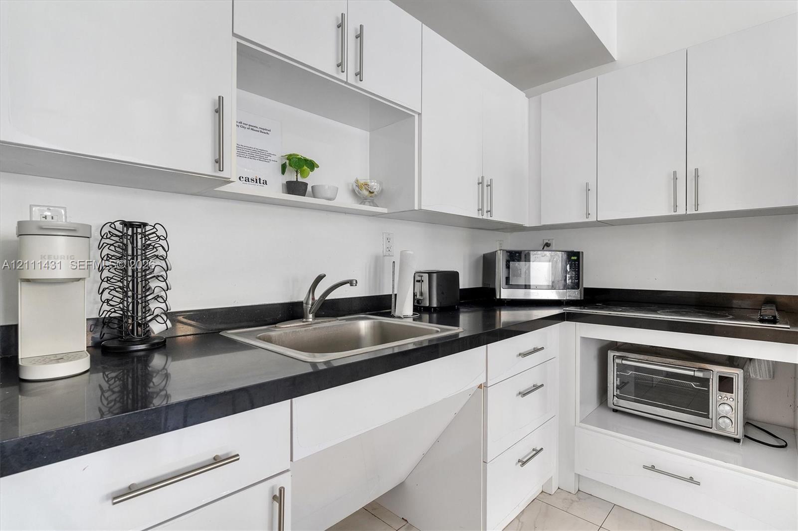 Kitchen featuring white flat-panel cabinetry, dark stone countertops, stainless steel sink with a high-arch faucet, and integrated appliance storage
