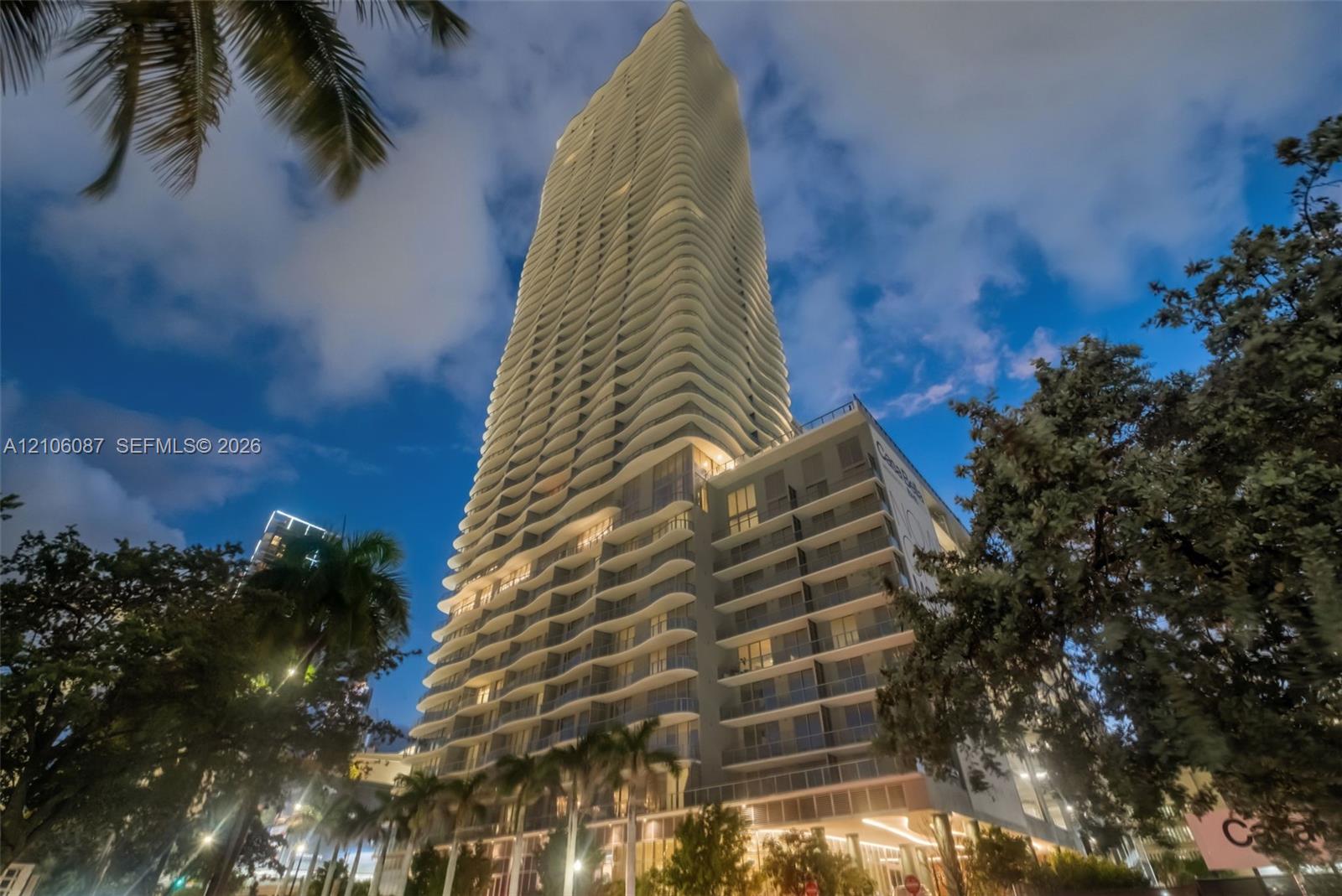 High-rise residential tower featuring a curvilinear architectural facade, extensive exterior balconies, glass curtain walls, and integrated ground-lev