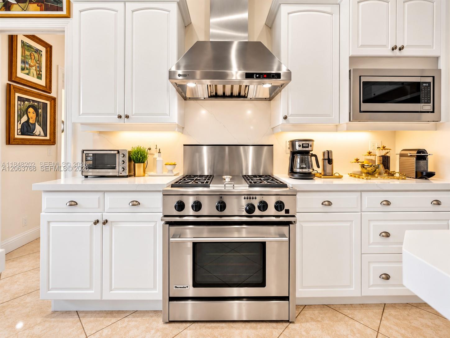 White cabinetry with metallic hardware, stainless steel gas range, professional-grade ventilation hood, built-in stainless steel microwave, and stone 