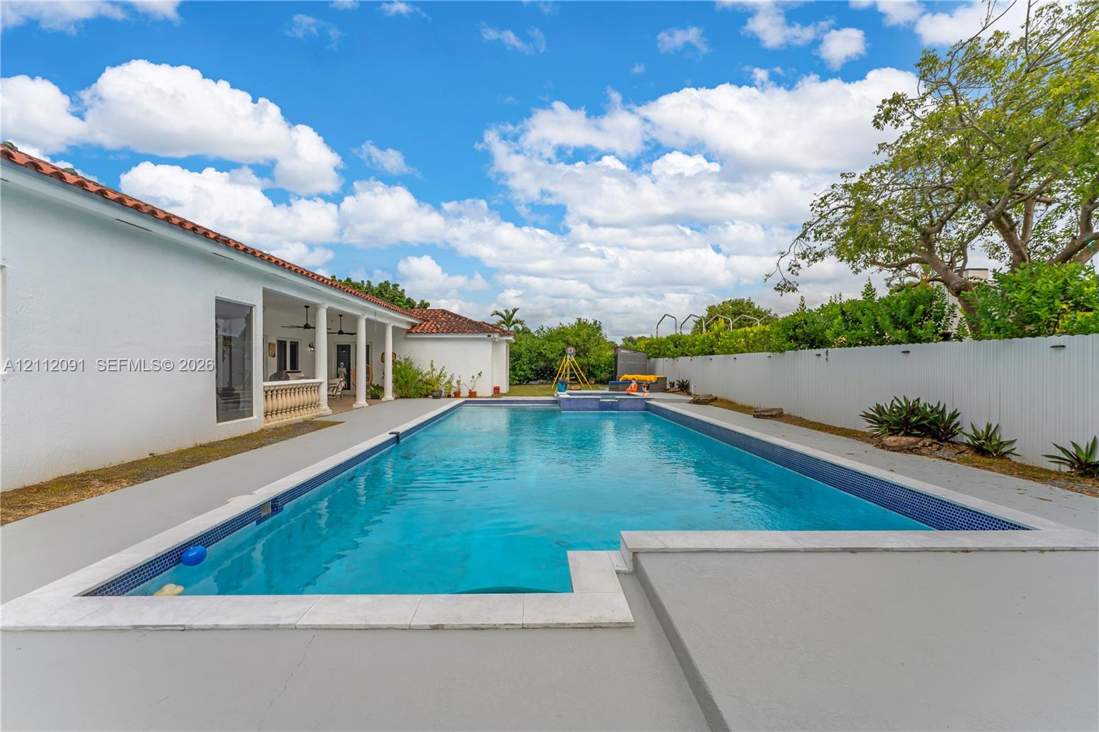 In-ground swimming pool with tile border, expansive concrete decking, covered patio with structural columns, and perimeter privacy fencing