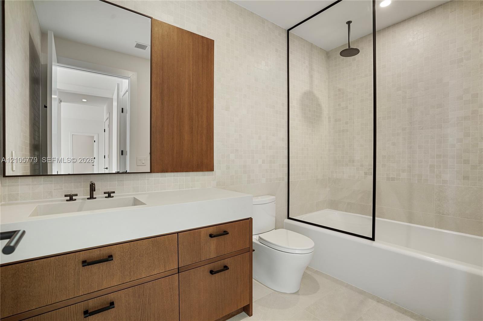Wood-finish vanity with a rectangular sink, stone-composite countertop, matte black hardware, rainfall shower head, and glass partition