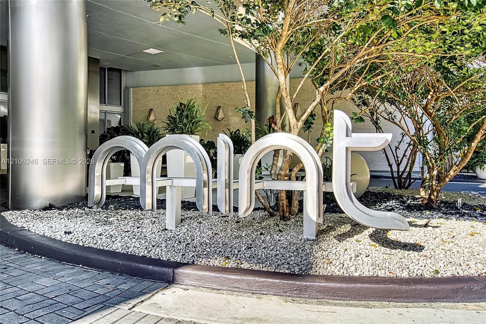 Exterior entrance featuring brushed metal structural columns, stone-textured facade wall, sculptural metal signage, and gravel landscaping with integr