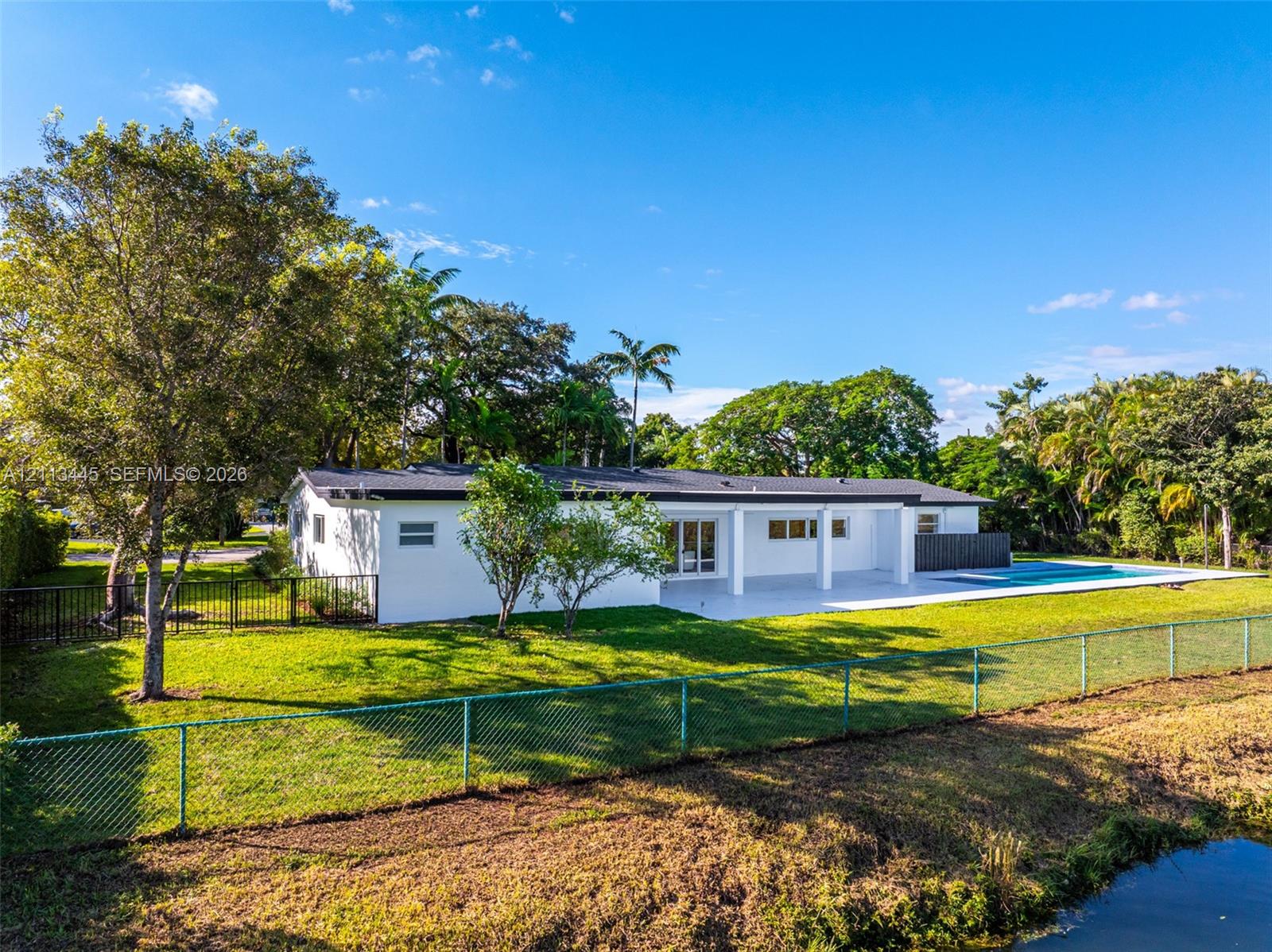 Exterior featuring a white stucco facade, covered patio, attached rectangular swimming pool, metal perimeter fencing, and mature landscaping