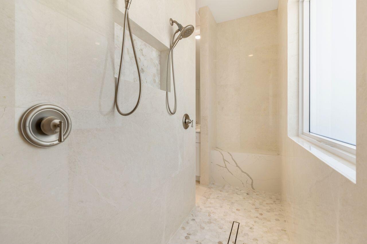 Spacious shower featuring light-toned tile walls, dual shower heads with brushed nickel fixtures, a built-in bench with veined stone, pebble-finish fl