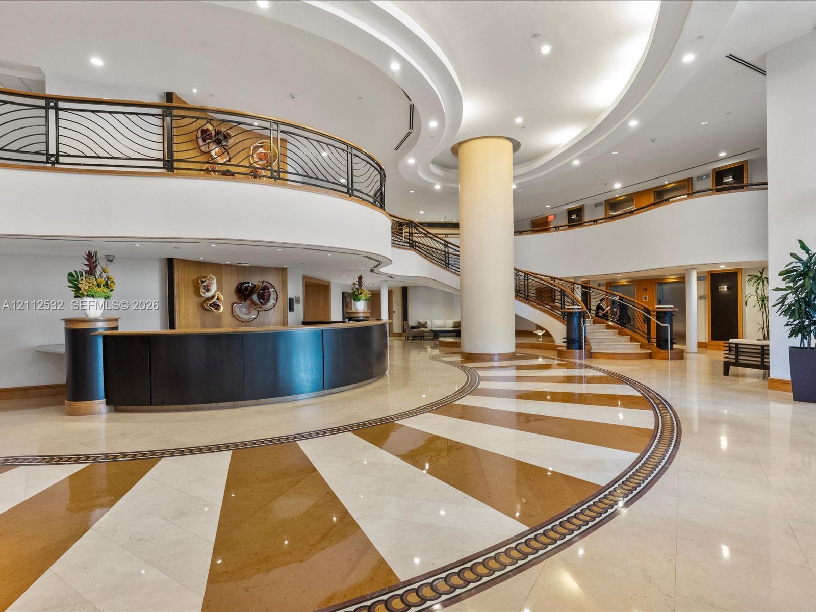 Grand lobby featuring expansive radial-patterned stone flooring, a curved reception desk with wood paneling, a sweeping staircase with ornate metal ra