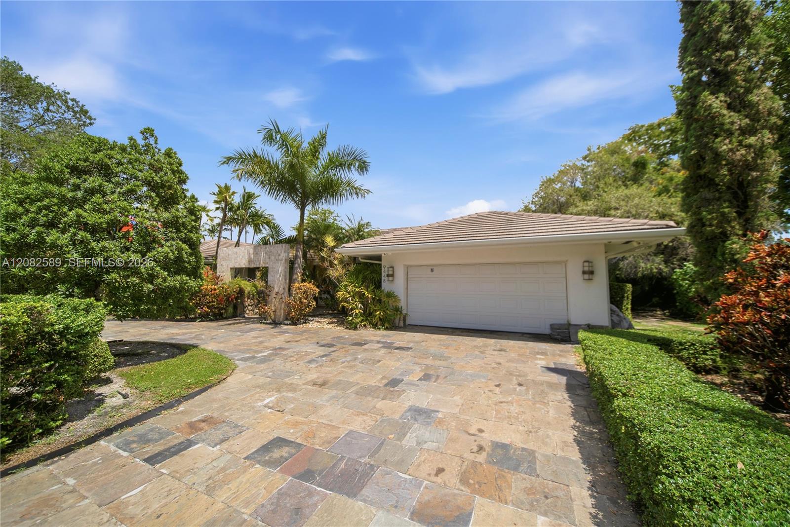 Spacious stone-paved driveway with an attached two-car garage, surrounded by lush tropical landscaping.