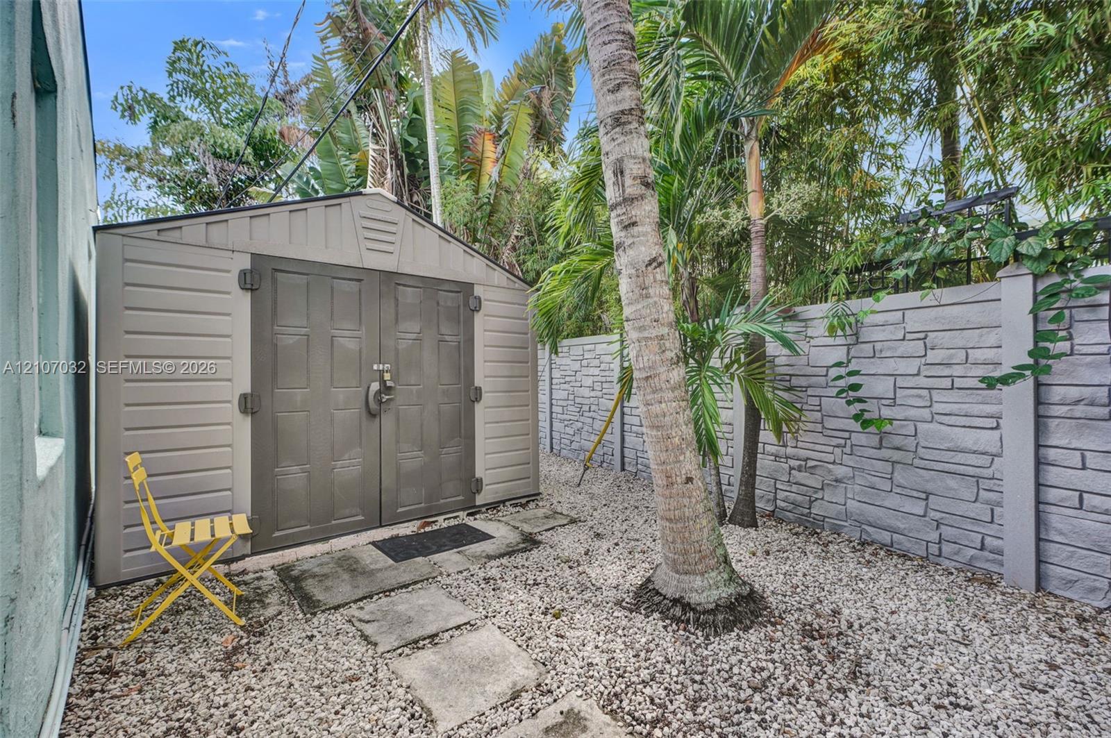 Exterior storage shed featuring dual-panel doors, accompanied by a stone-pattern privacy wall, stone paver pathway, and gravel ground cover
