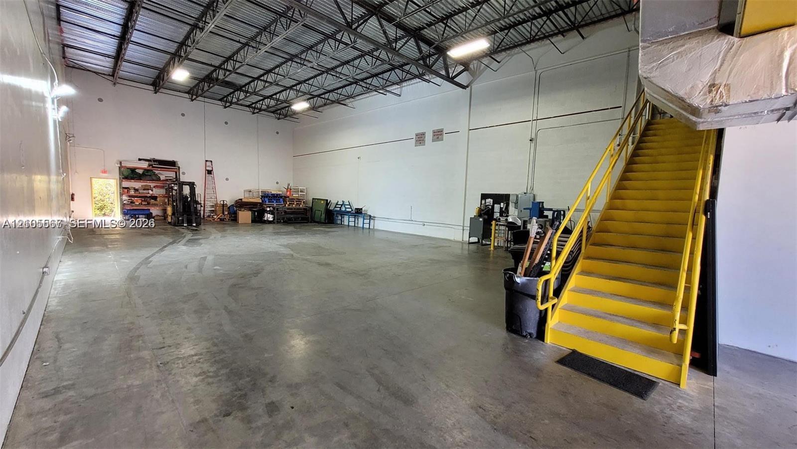 Expansive concrete flooring with a high exposed ceiling, ample overhead lighting, and a prominent yellow staircase with handrails