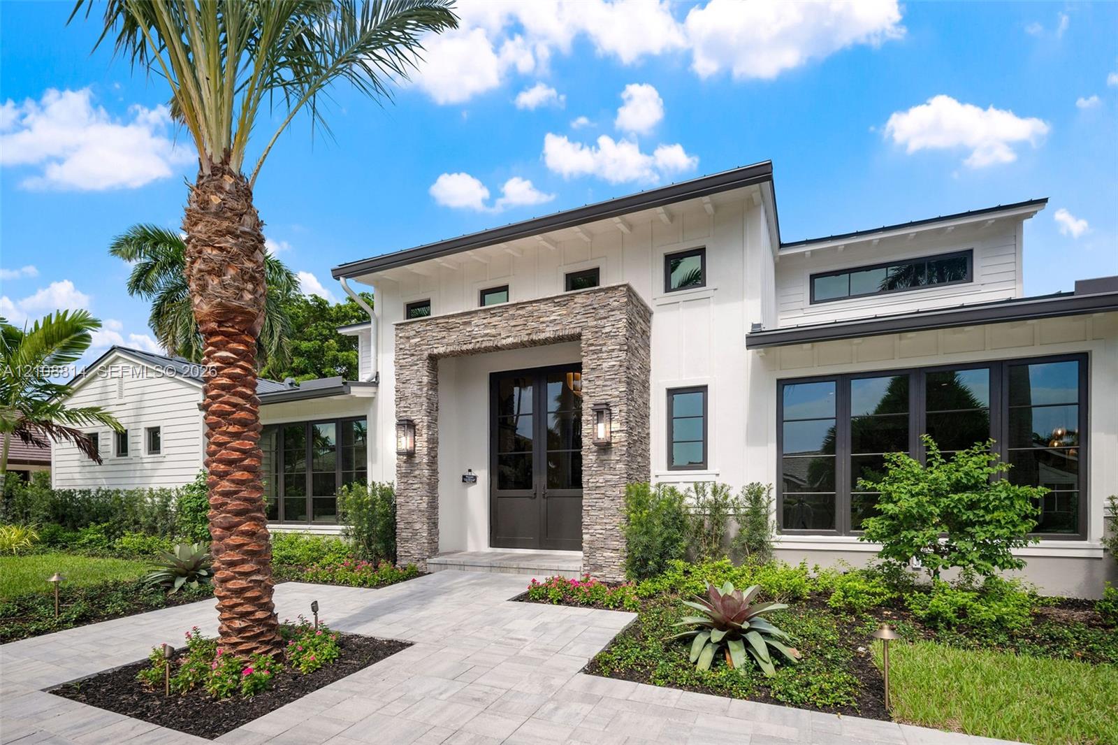 Modern exterior featuring white vertical siding, a stone veneer entryway surround, metal roof accents, expansive dark-framed windows, and a paved walk