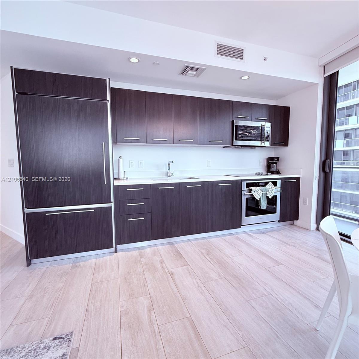 Kitchen featuring dark-finish cabinetry, light-toned stone-surface countertops, wood-finish flooring, and integrated stainless-steel appliances