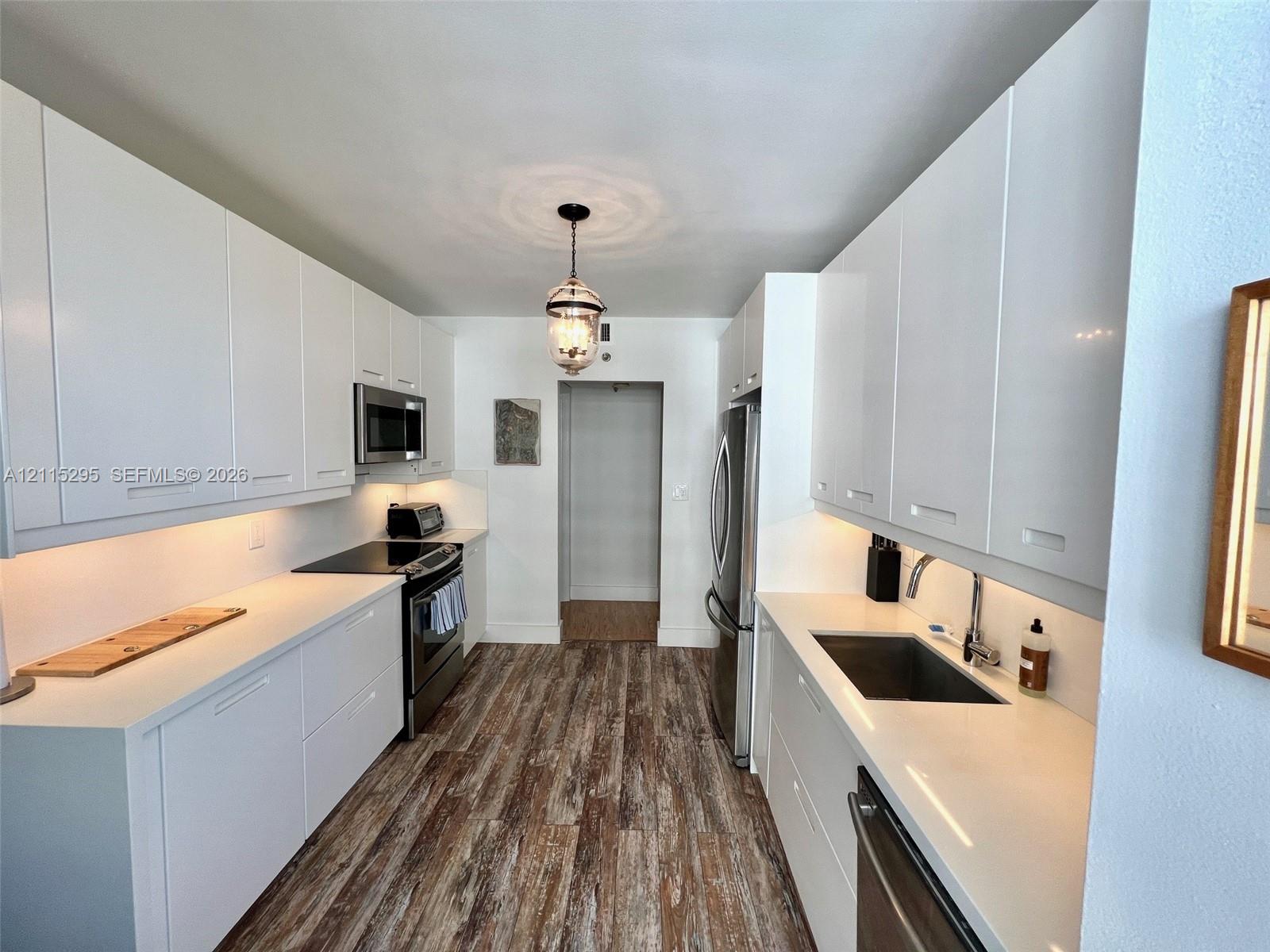 Galley kitchen featuring flat-panel white cabinetry, solid-surface countertops, stainless steel appliances, under-cabinet lighting, and wood-finish fl