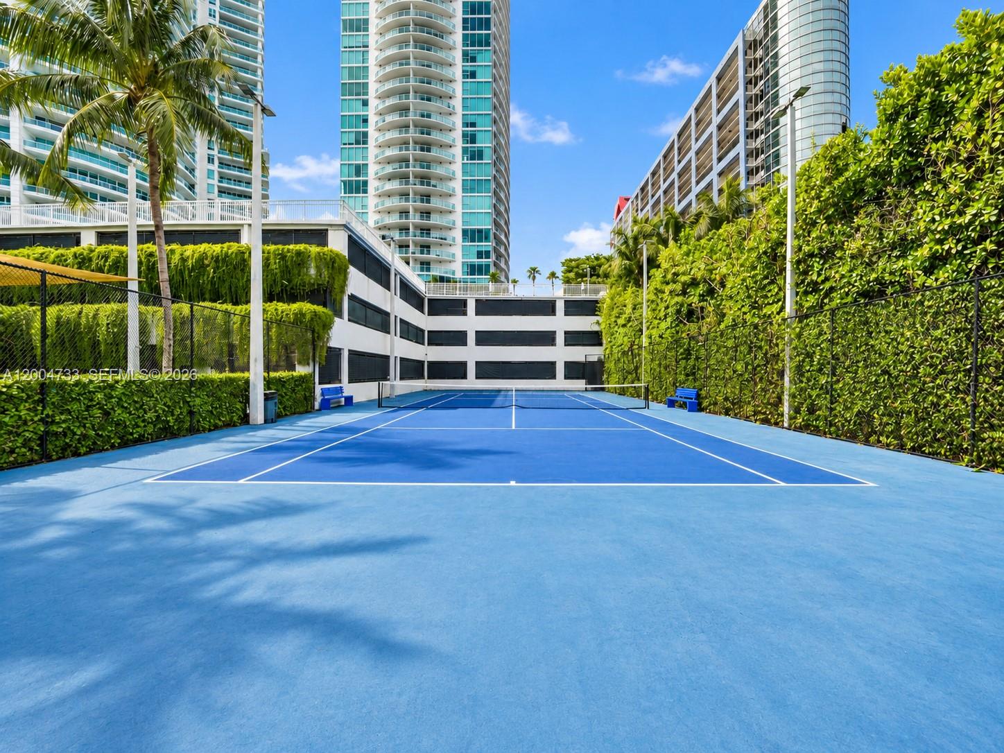 Hard-court tennis facility featuring blue acrylic surfacing, black perimeter chain-link fencing, integrated pole-mounted lighting, and extensive perim