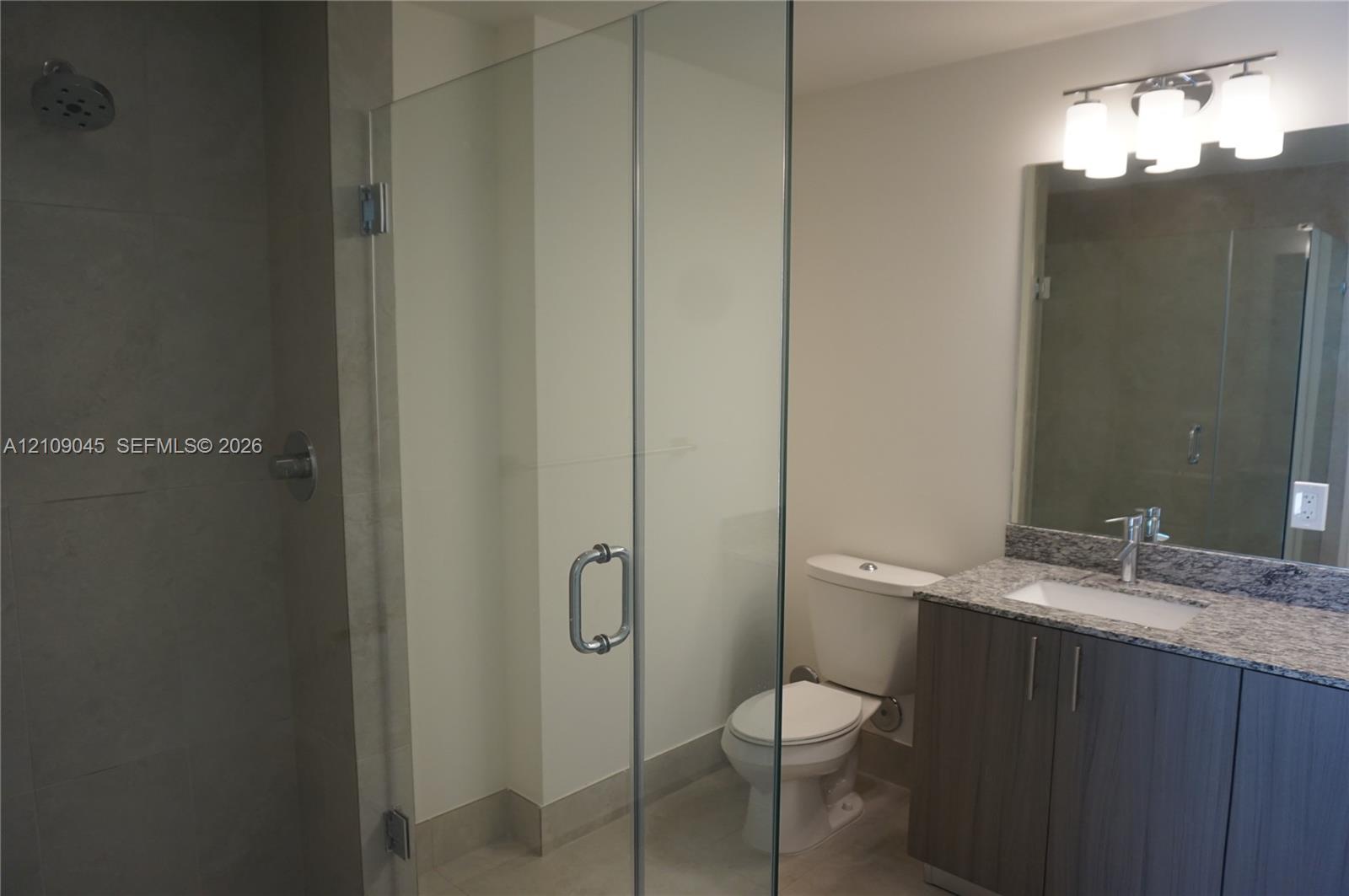 Bathroom featuring a walk-in shower with a frameless glass enclosure, vanity with stone-patterned countertop, under-mount sink, and multi-bulb integra