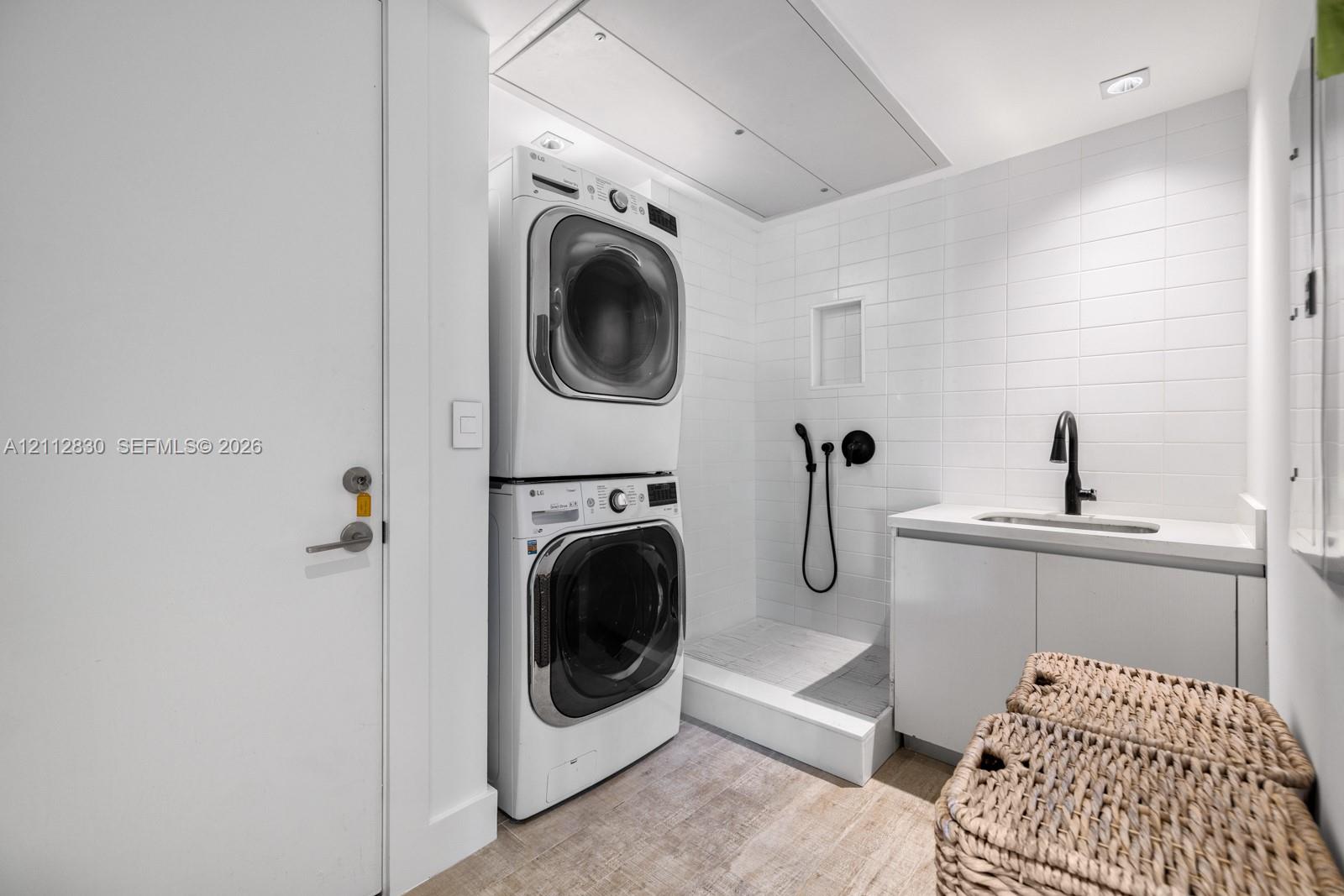 Laundry area featuring stacked appliances, tiled shower enclosure with handheld sprayer, built-in cabinetry, solid-surface countertop, and matte black