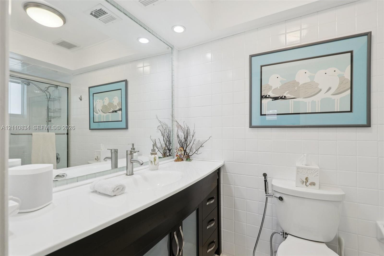 Bathroom vanity featuring a solid surface countertop, integrated sink, dark-toned cabinetry, full-width mirror, and shower enclosure with a glass door
