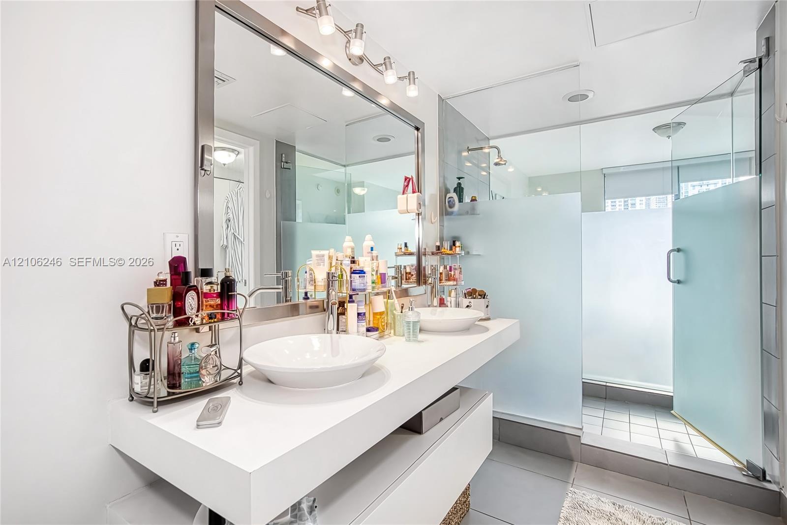 Floating dual-vanity with rectangular vessel sinks, expansive wall-to-wall mirror, overhead track lighting, and a walk-in shower enclosed by frosted g