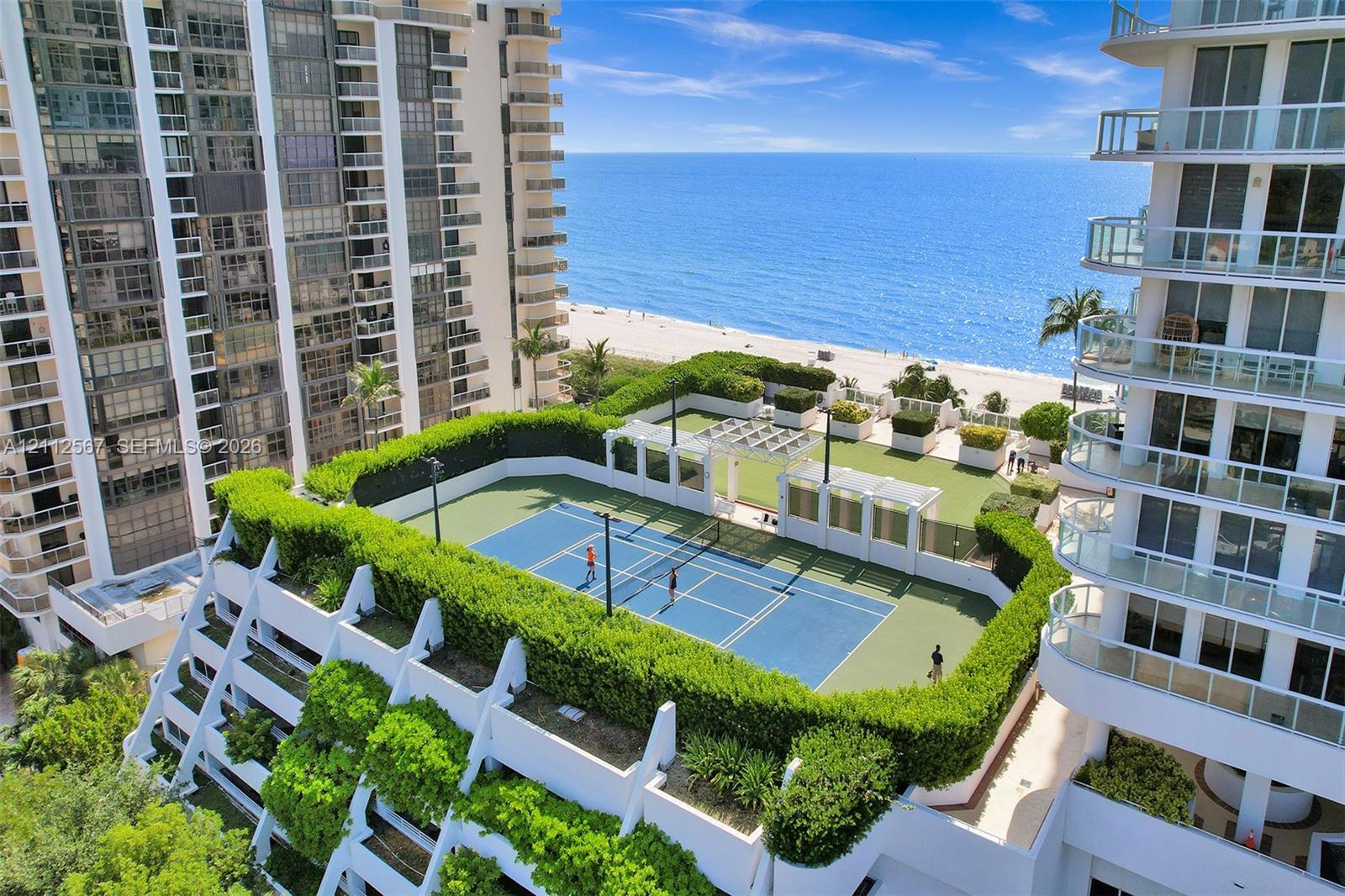 Hard-surface tennis court with perimeter landscaping, pergola-covered seating area, and synthetic turf lawn