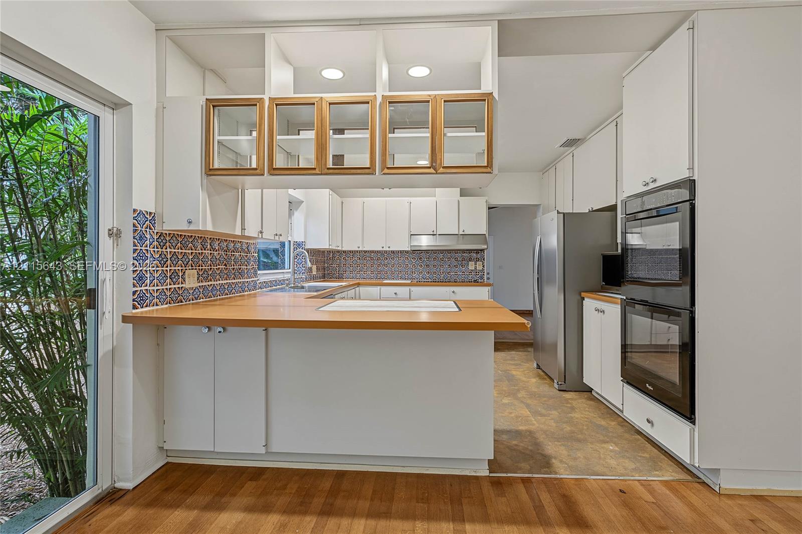 Kitchen featuring white cabinetry with glass-front uppers, wood-finish countertops, patterned tile backsplash, and built-in double wall ovens