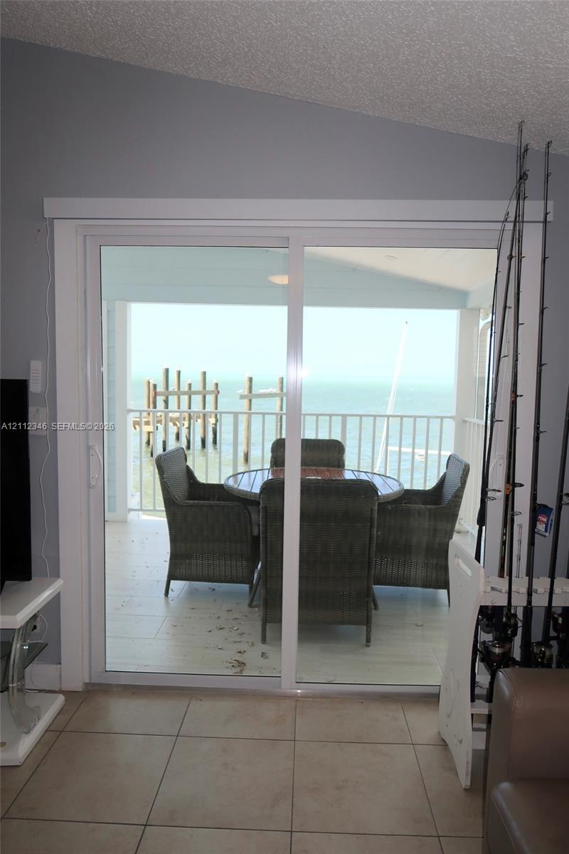 Sliding glass door opening to a covered balcony with a white metal railing, tile flooring, and coastal water backdrop
